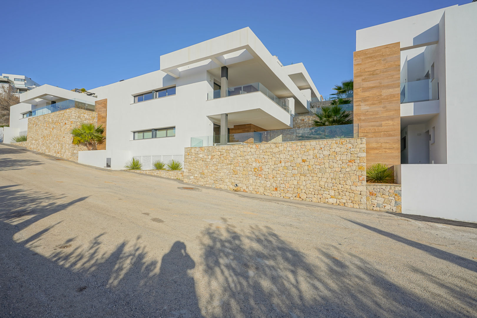 Modern facade at Monte Cala Gardens featuring natural stone walls, spacious glass-railed terraces, and wood-effect accents under a clear blue sky.