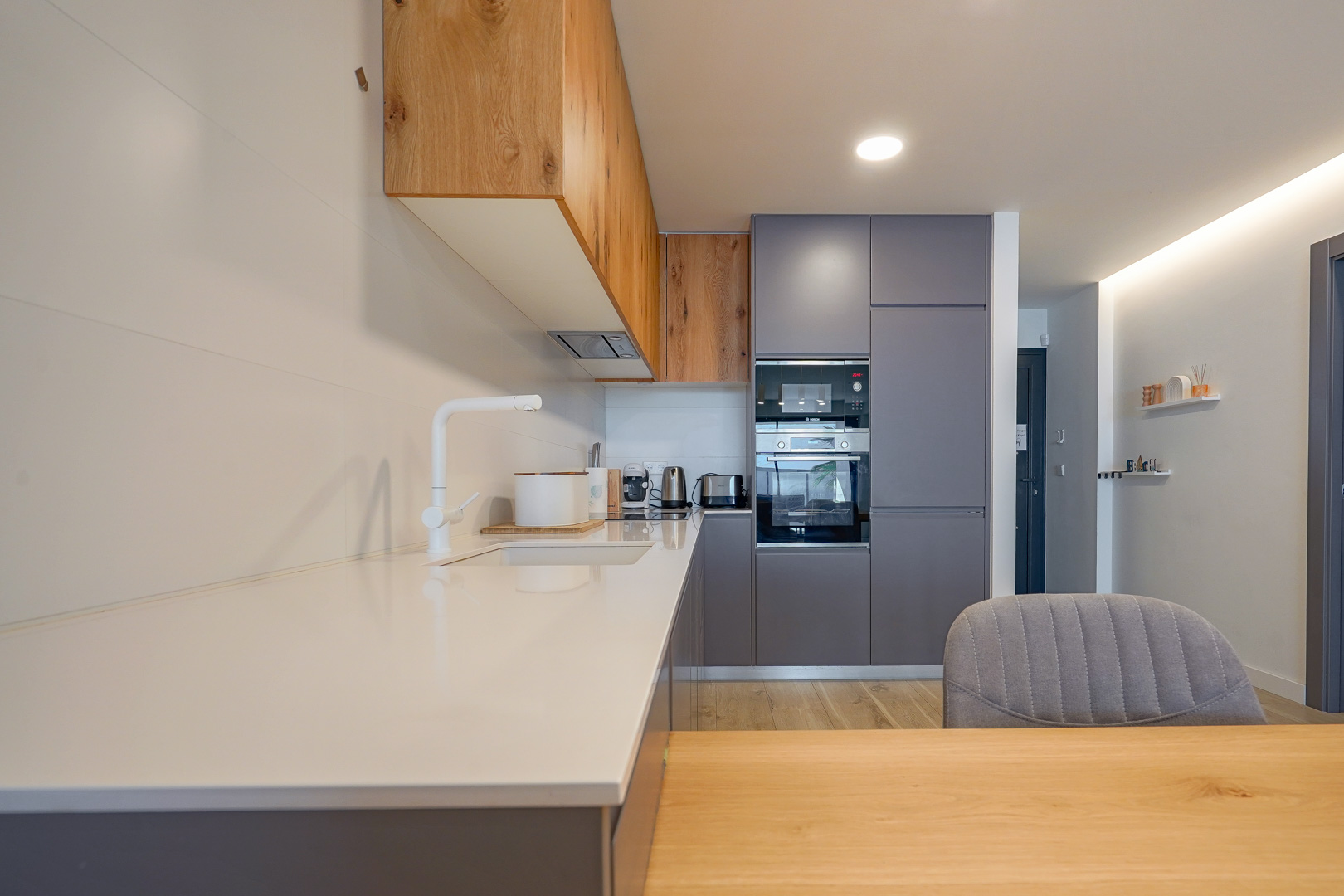 Modern kitchen featuring a white countertop, grey and wood cabinetry, integrated appliances, and a minimalist white faucet under recessed lighting.