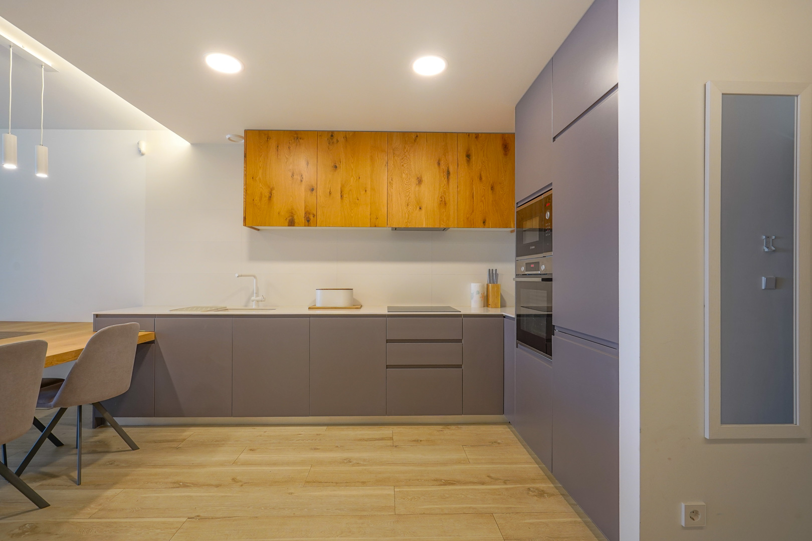 Modern minimalist kitchen featuring matte grey and natural wood cabinetry, integrated appliances, a slim white countertop, and light wood-effect flooring.