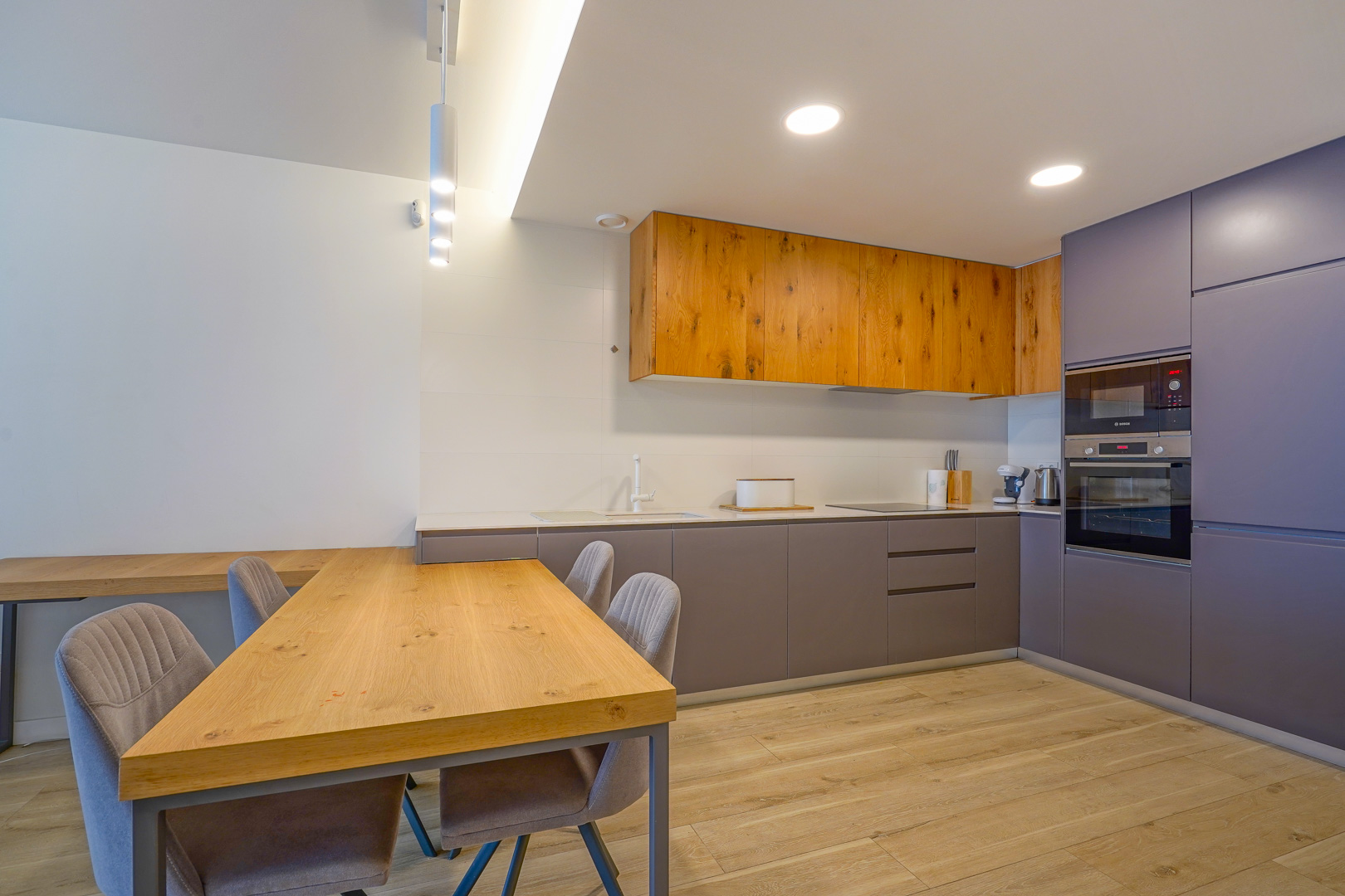 Modern open-plan kitchen featuring natural wood and grey cabinetry, integrated appliances, and a matching wooden dining table with upholstered chairs.