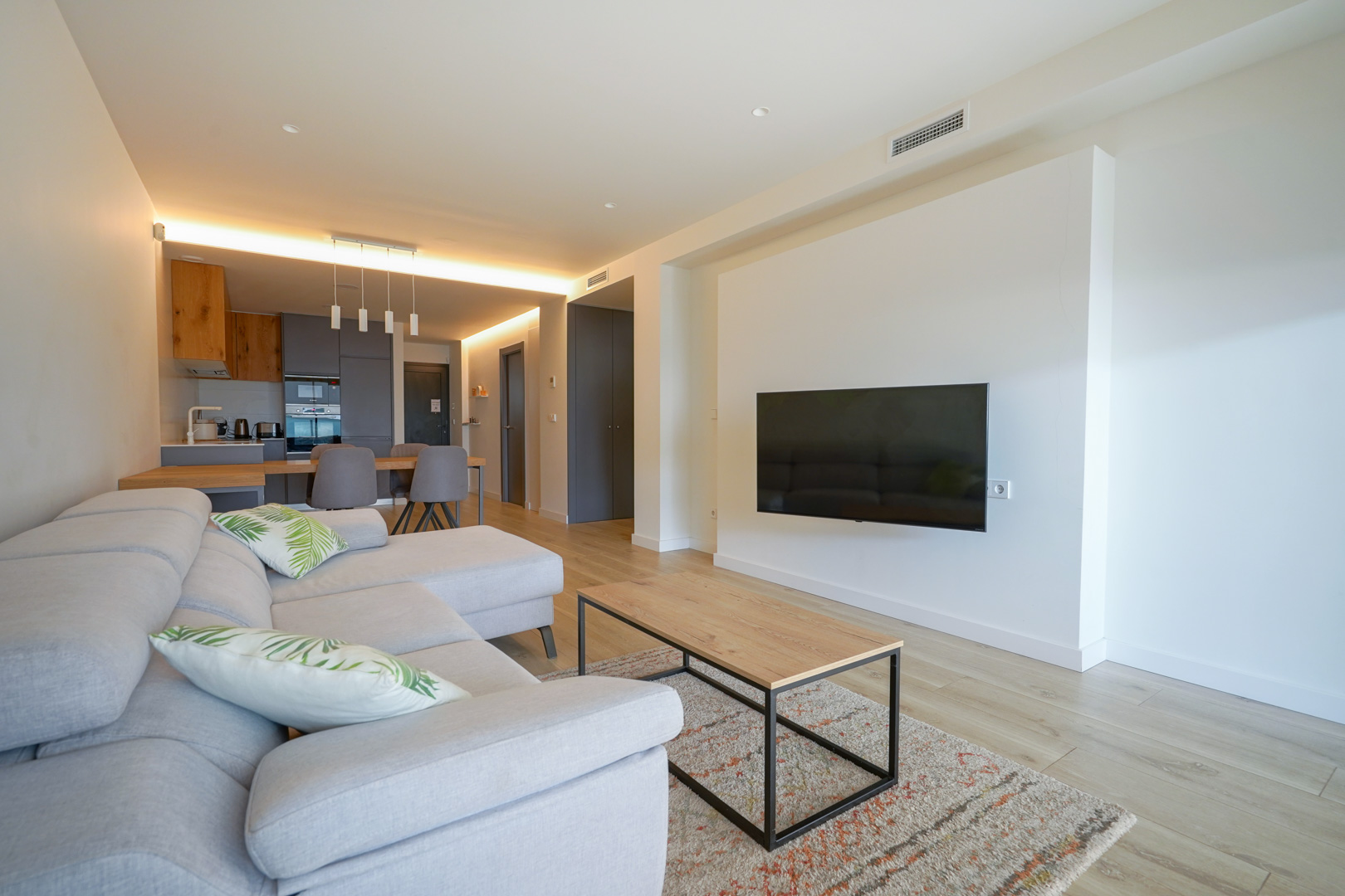 Open-plan living area featuring a wall-mounted television, light wood flooring, and an integrated modern kitchen with recessed LED lighting.