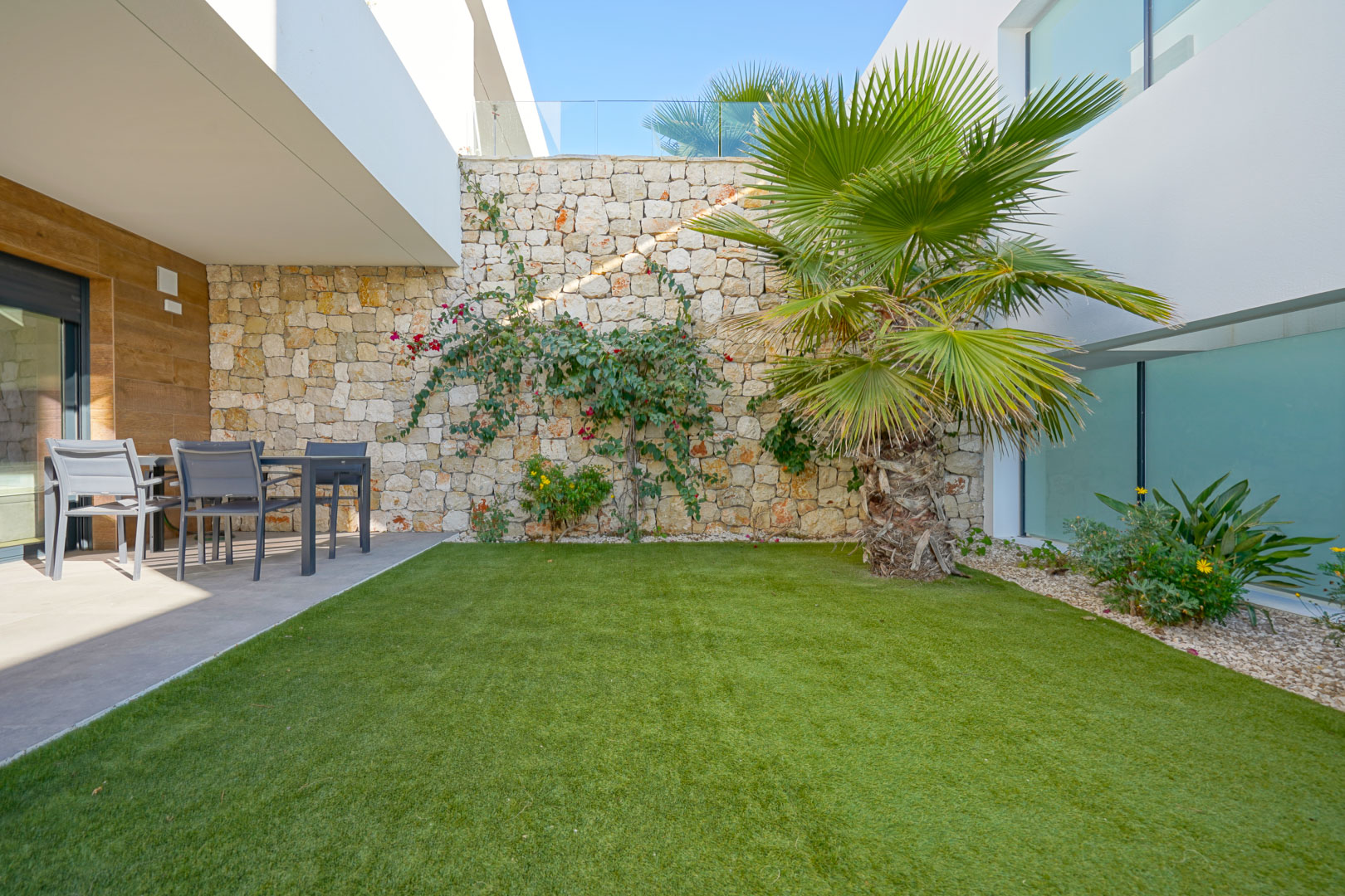 Private garden featuring artificial turf, a palm tree, and a natural stone wall. A covered terrace area provides a shaded outdoor dining space with modern finishes.