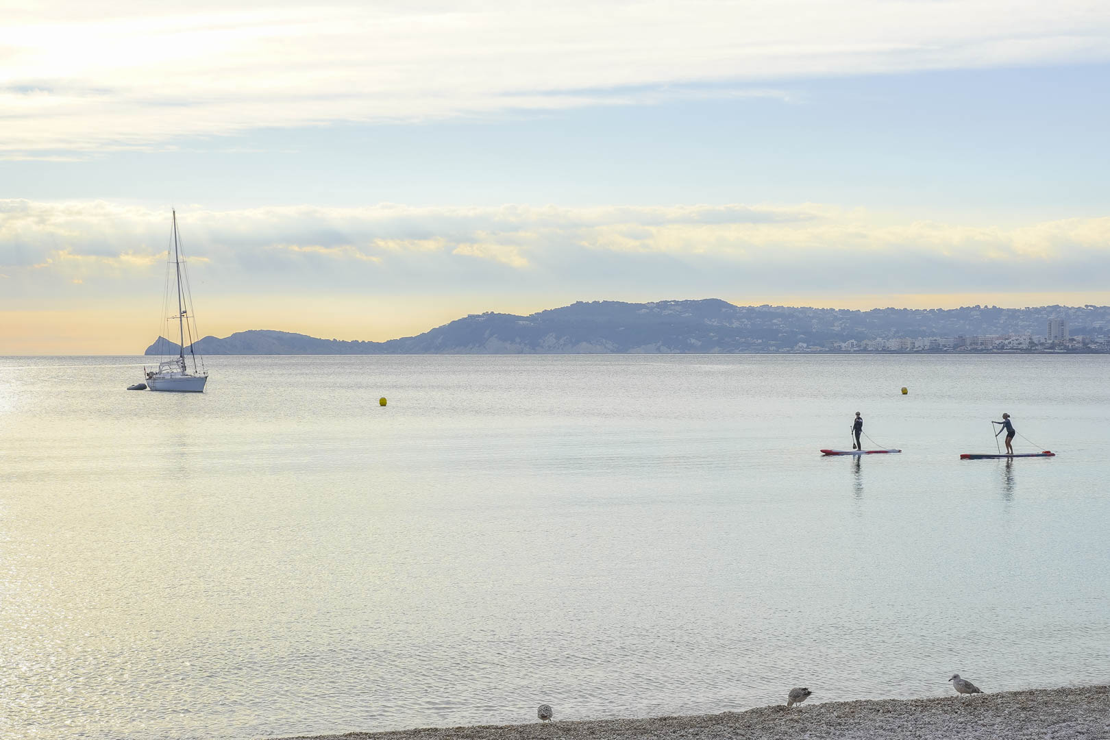 Panoramic view of Jávea's bay featuring calm Mediterranean waters, an anchored sailboat, and paddleboarders near the pebble beach of Cala Blanca.