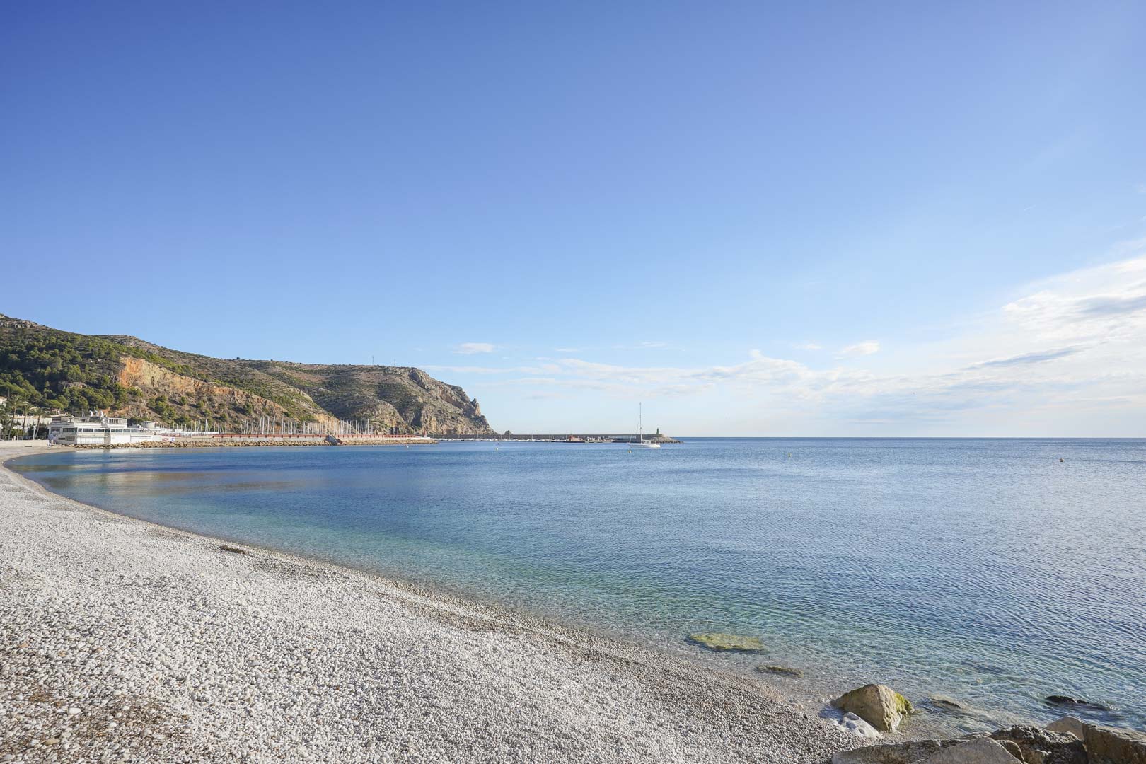 Panoramic view of the Jávea coastline featuring a white pebble beach, turquoise waters, and the local marina set against the rugged cliffs.