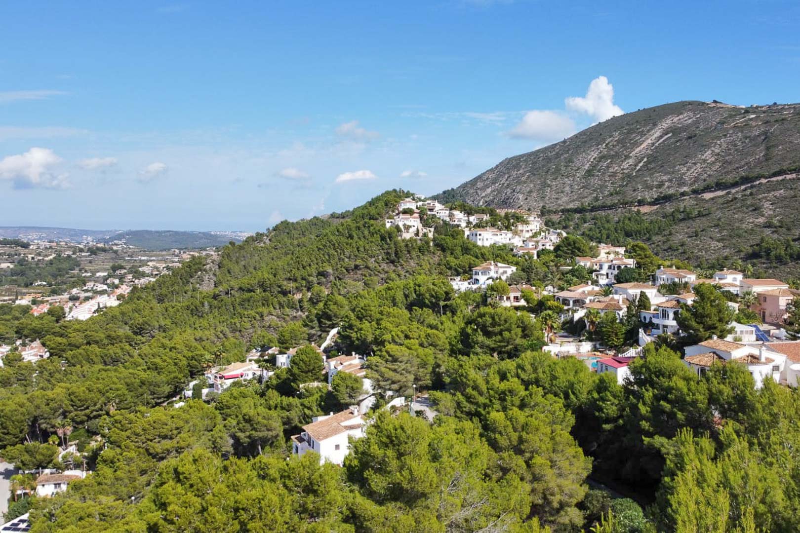 Aerial view of the Paichi residential area in Moraira, showing white villas nestled within a dense pine forest with expansive mountain backdrops.
