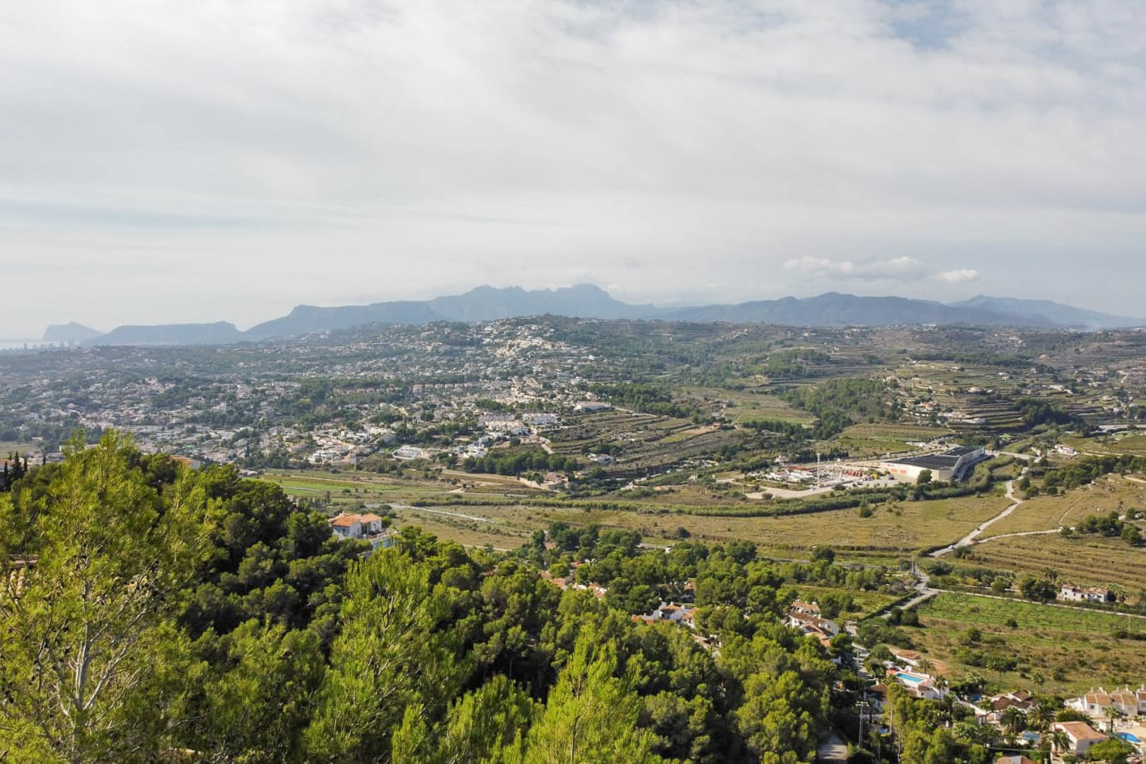 Elevated panoramic view from the plot overlooking the Moraira valley, featuring Mediterranean pine forests and distant mountain ranges.
