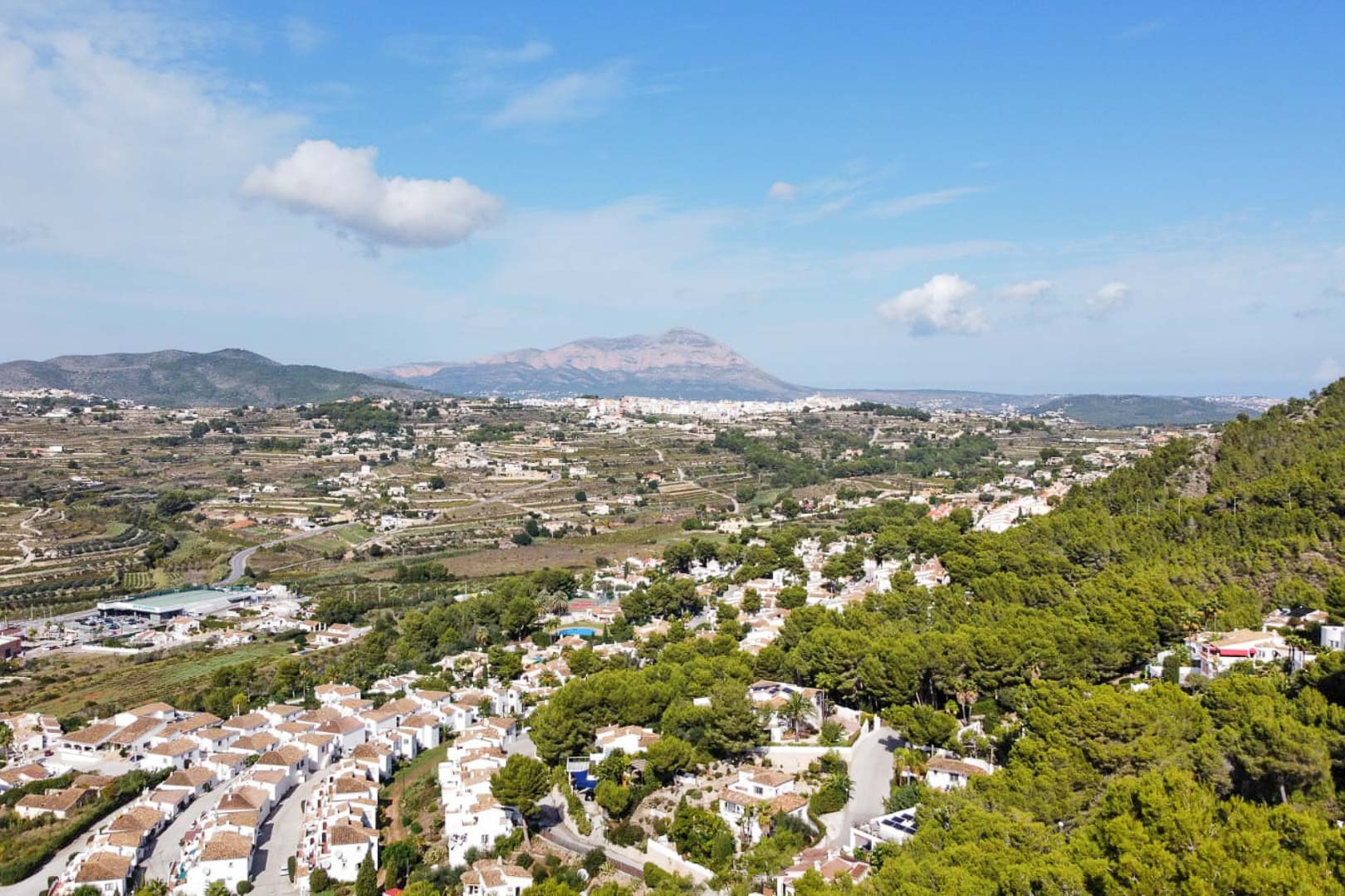 Elevated panoramic view showcasing Mediterranean pine forests and white-walled villas, featuring a prominent mountain silhouette on the horizon under a clear sky.