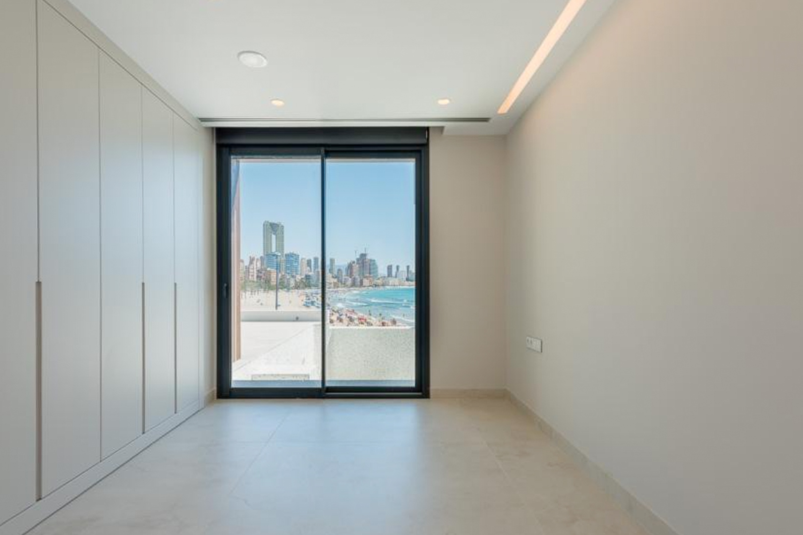 Bedroom with floor-to-ceiling wardrobes, integrated LED lighting, and a sliding glass door showcasing views of Poniente beach and the city skyline.