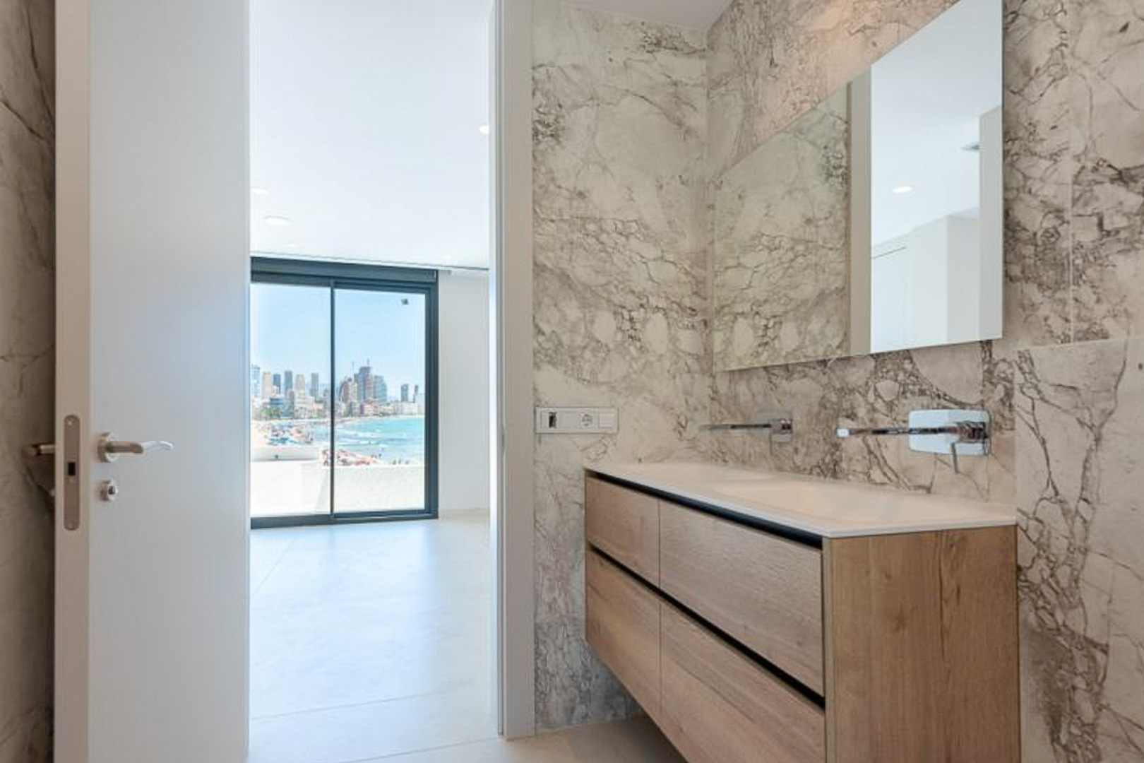 Designer bathroom featuring marble-effect walls, a floating oak-finish double vanity, and integrated faucets, offering views toward the coastline.