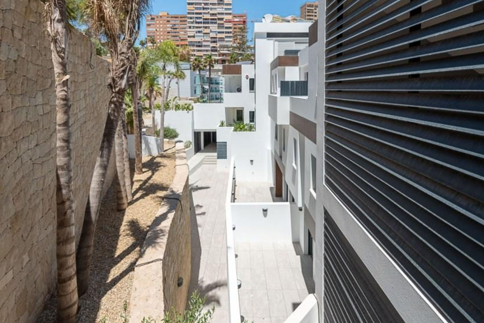 Exterior view of the complex featuring white facades, a natural stone wall, and palm trees. Modern horizontal louvers and paved walkways define the architecture.