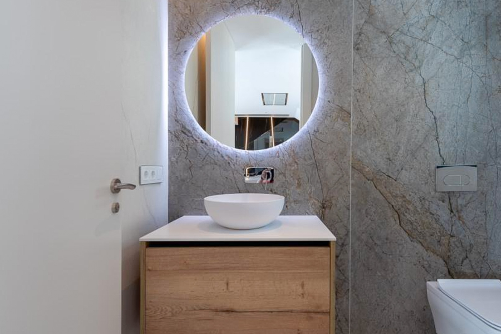Guest bathroom featuring grey marble-effect wall tiles, a circular backlit mirror, and a wooden vanity with a white vessel sink and wall-mounted faucet.