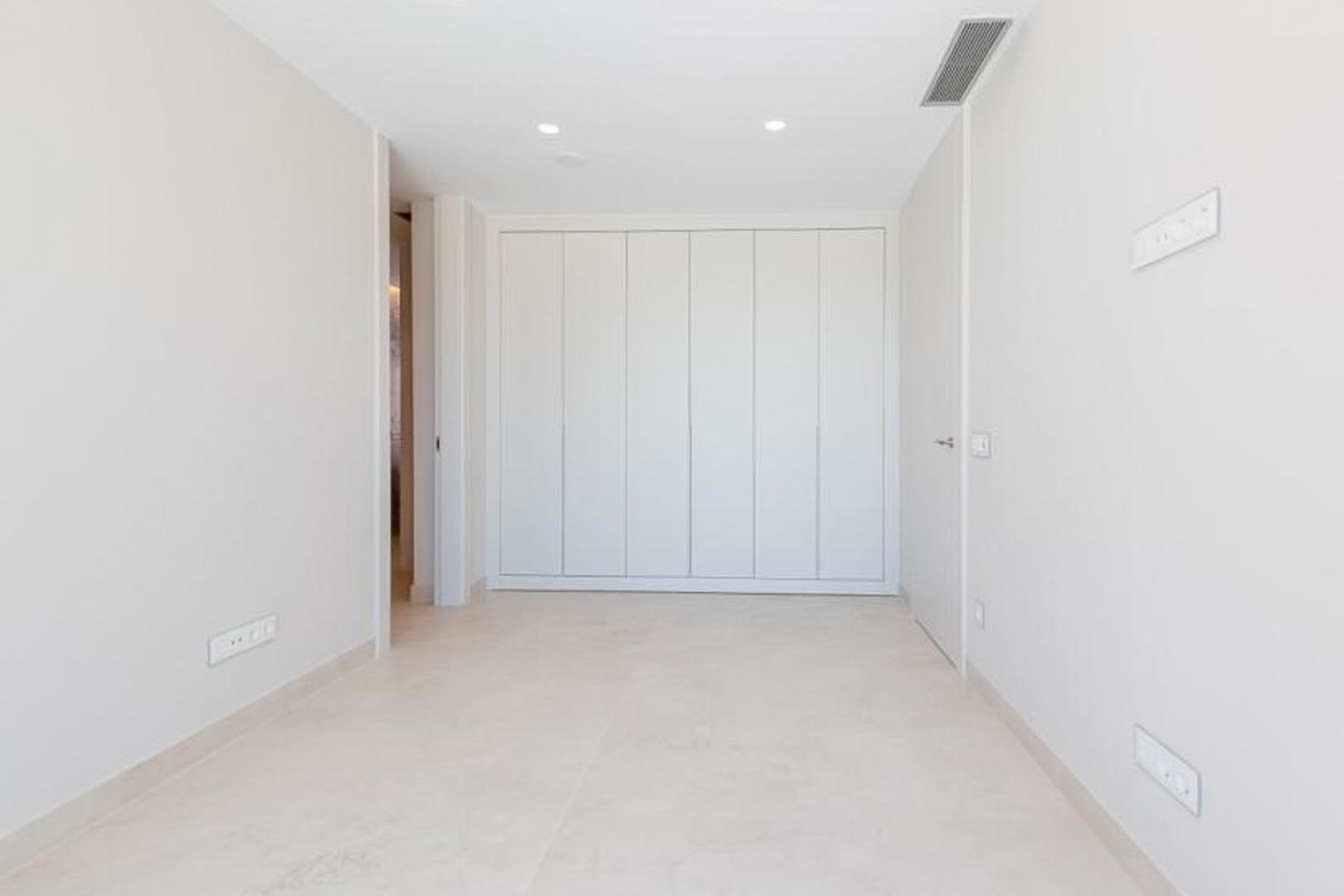 Minimalist bedroom featuring floor-to-ceiling built-in wardrobes, light porcelain flooring, and recessed ceiling lighting with integrated air conditioning vents.