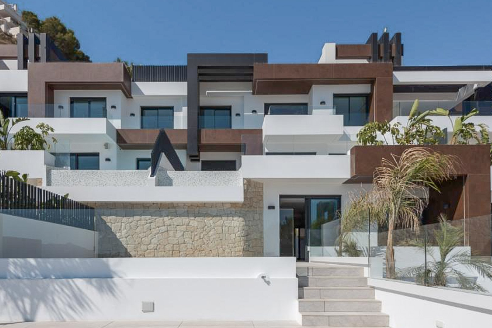 Modern architectural facade featuring white and brown geometric volumes, natural stone cladding, glass railings, and tiered terraces with integrated landscaping.
