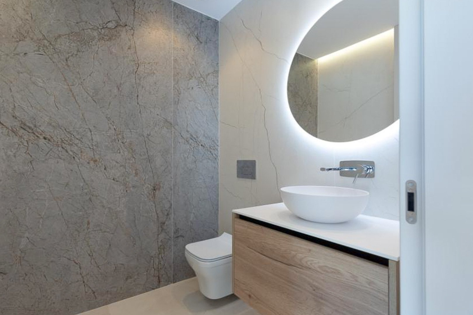 Modern bathroom featuring a floating wooden vanity, vessel sink, and a circular backlit mirror against large-format marble-effect wall tiles.