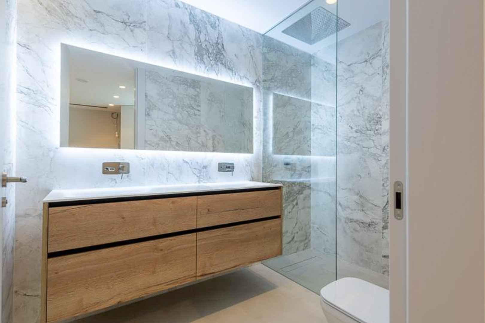 Modern bathroom featuring marble-effect tiling, a floating wooden double vanity, and a backlit mirror. Includes a walk-in shower with a glass partition.
