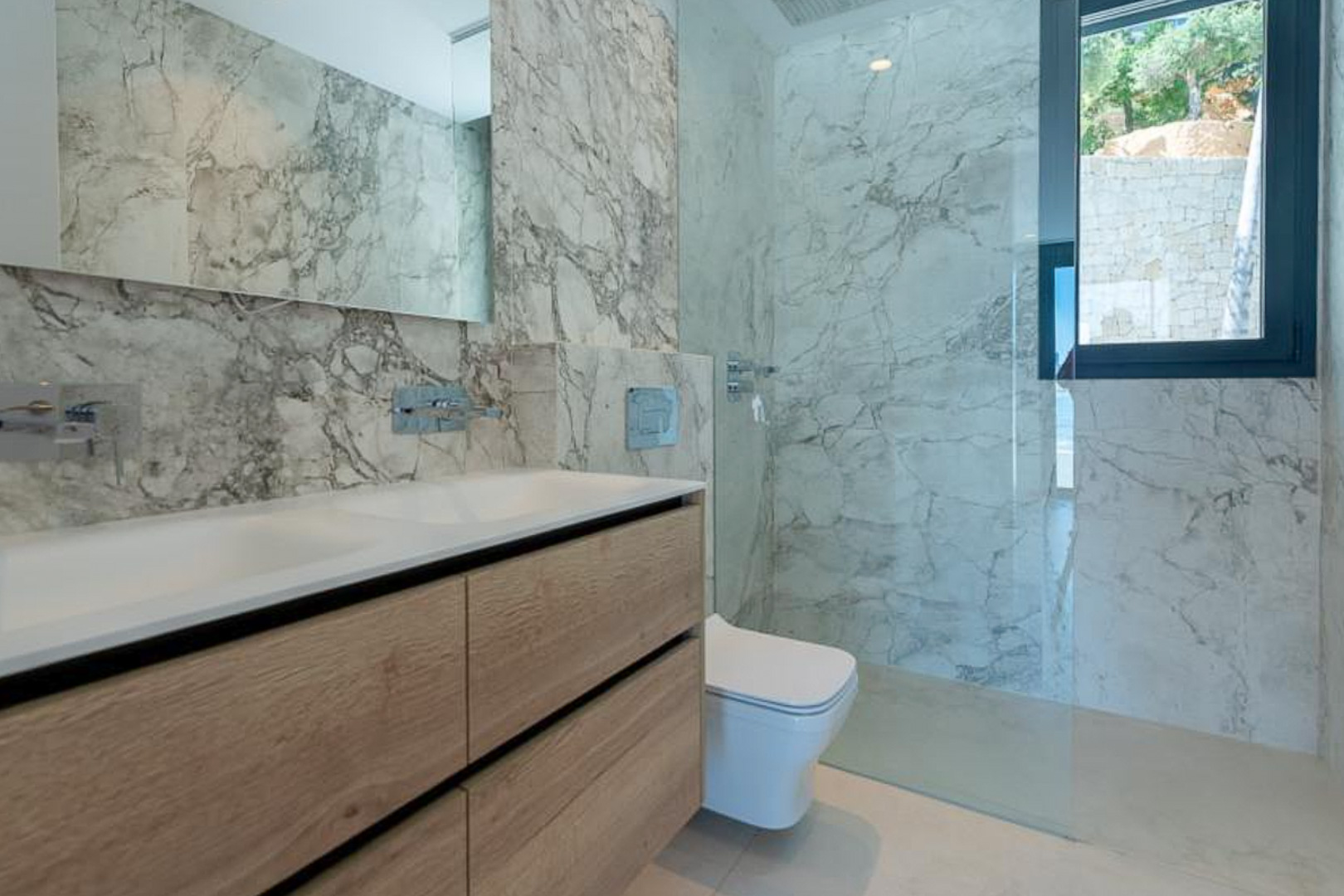 Modern bathroom featuring marble-effect wall tiling, a light wood double vanity, and a walk-in shower with a frameless glass partition.