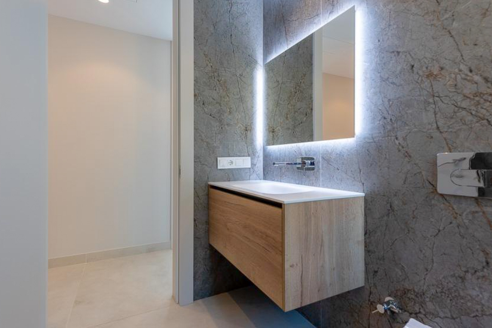 Modern guest bathroom with a floating wooden vanity, integrated sink, and backlit mirror against grey marble-effect wall tiles.