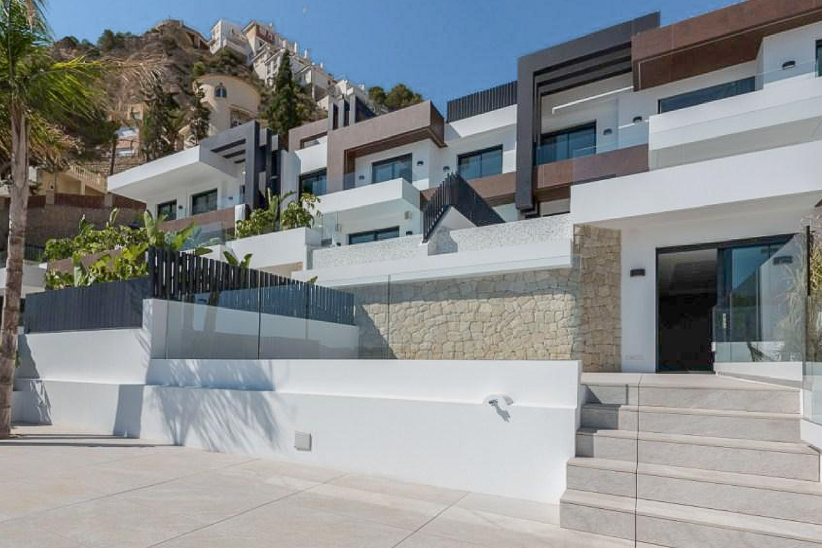 Modern tiered facade featuring natural stone walls, expansive terraces with glass railings, and porcelain tile stairs under a clear Mediterranean sky.