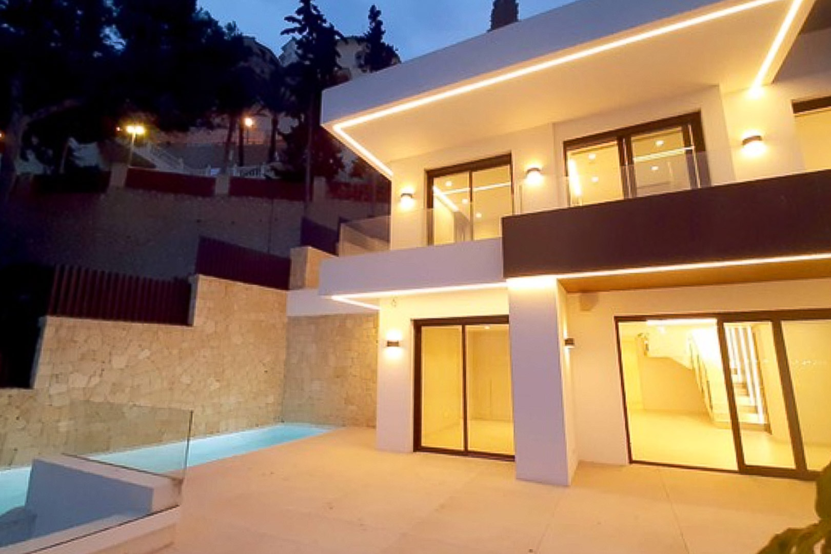 Modern two-story facade featuring integrated LED lighting, large sliding glass doors, and a private terrace with a pool set against a natural stone wall.