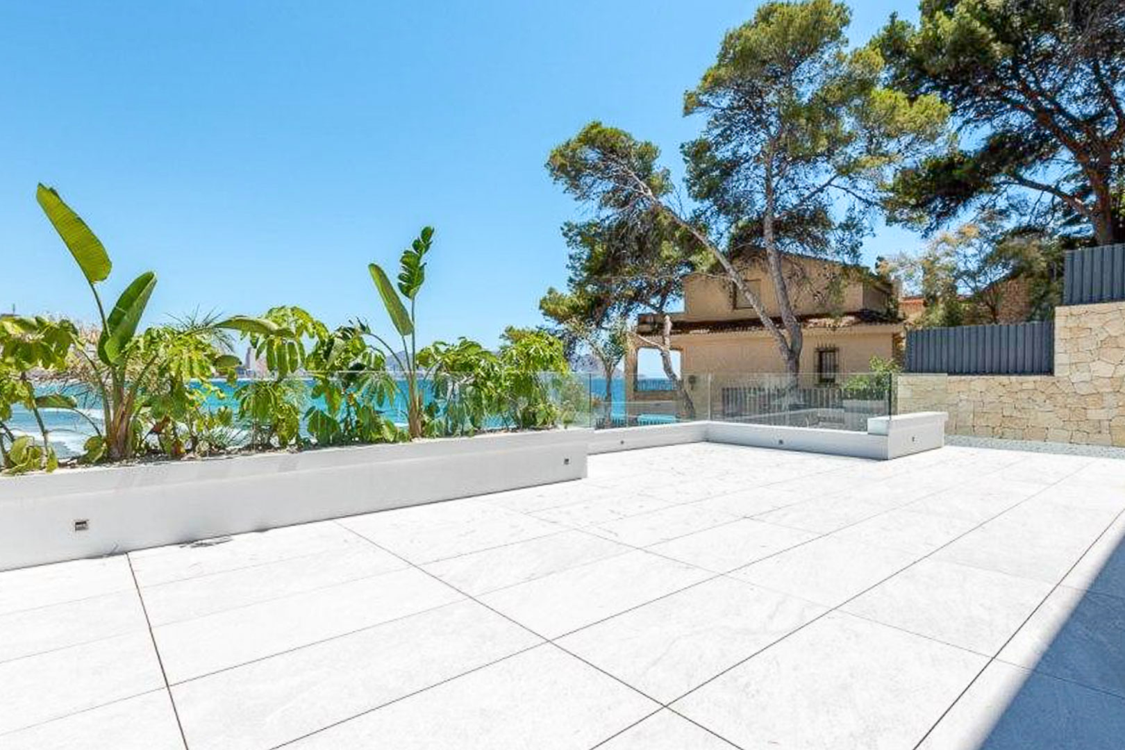 Outdoor terrace featuring large-format porcelain tiling, frameless glass railings, and integrated planters with tropical vegetation overlooking the Mediterranean Sea.