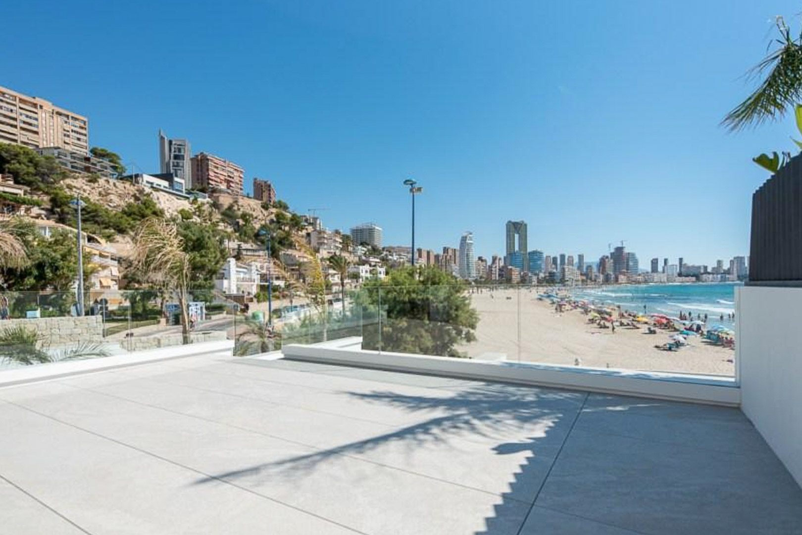 Outdoor terrace featuring large-format stone tiling and glass balustrades, overlooking the sandy beach and the Benidorm city skyline.