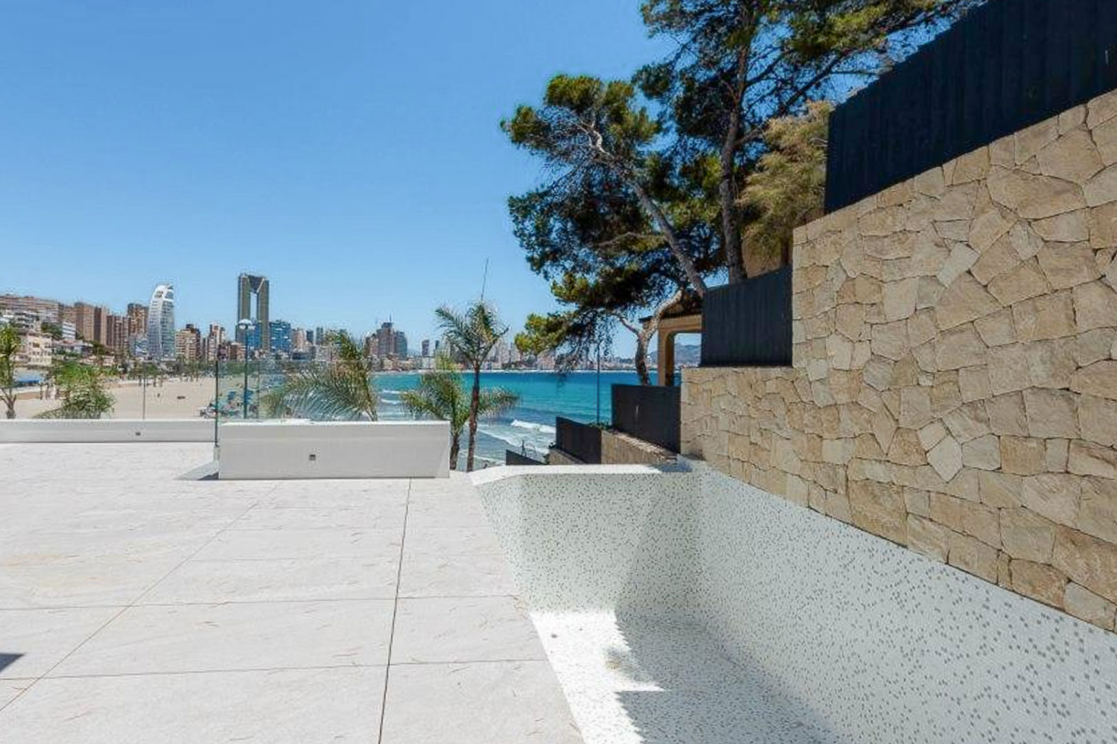 Outdoor terrace featuring light stone paving and a white mosaic pool, overlooking the Mediterranean Sea and Benidorm skyline beside a natural stone wall.