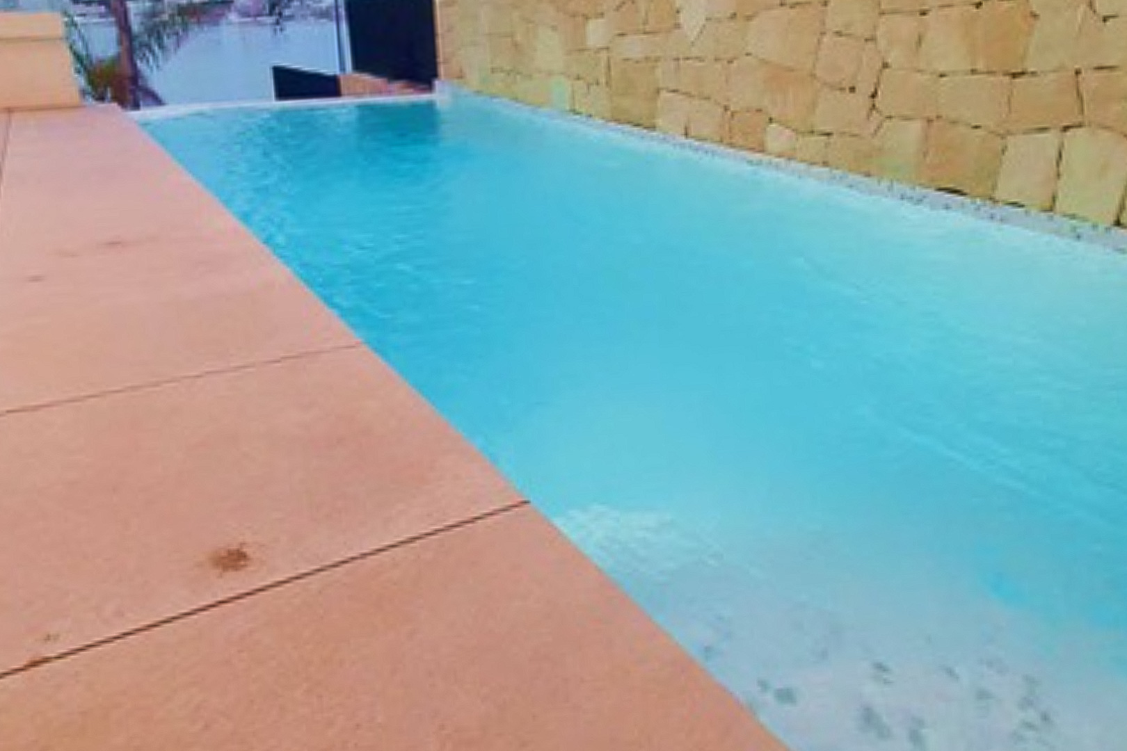 Private infinity pool featuring turquoise water, bordered by a terracotta-toned terrace and a rustic natural stone wall with sea views.