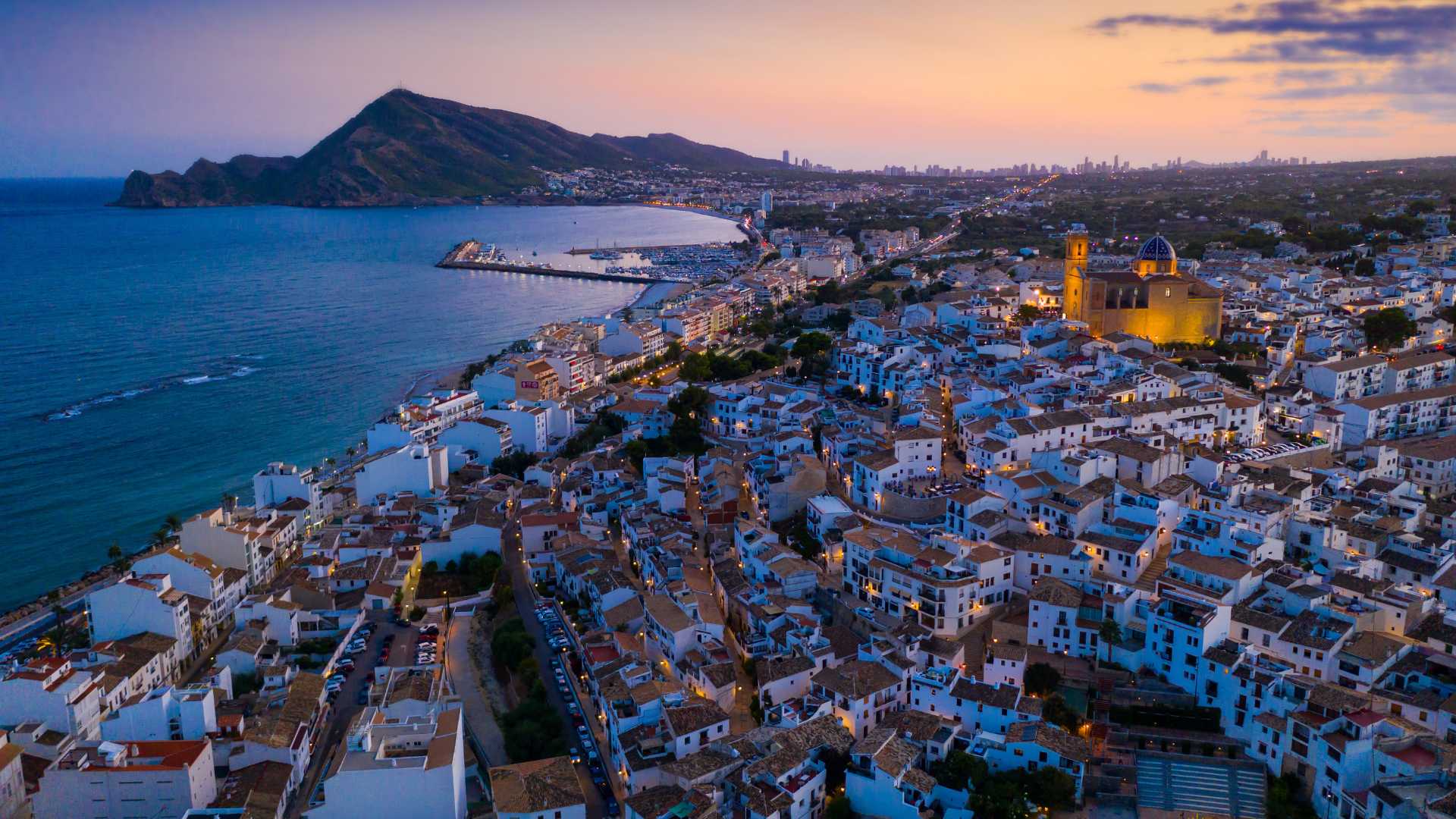 Aerial twilight view of Altea's old town, featuring the iconic blue-domed church, traditional white-washed architecture, and the Mediterranean coastline extending toward the horizon.