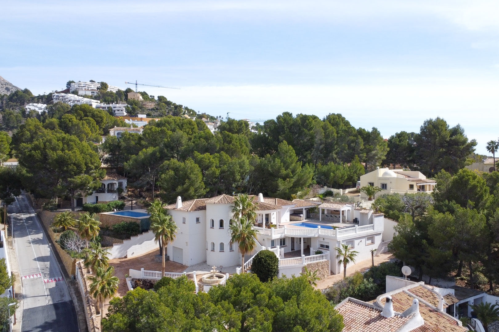 Aerial view of the Mediterranean villa featuring a white facade, circular tower, and elevated pool terrace nestled among lush pine trees in Altea.