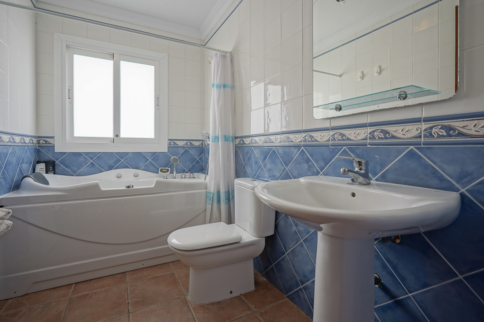 Bathroom featuring a whirlpool bathtub, pedestal sink, and classic blue and white wall tiling. Includes terracotta flooring and a frosted window for privacy.