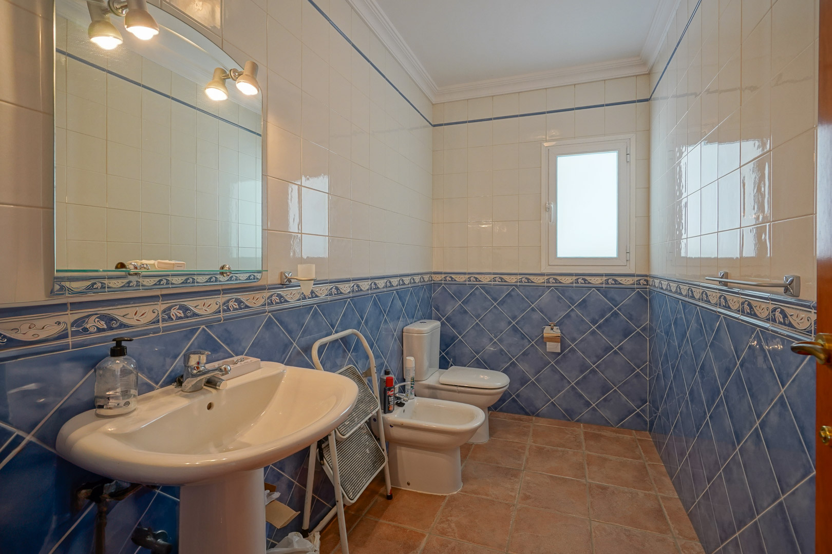 Bathroom featuring blue and white wall tiling with decorative borders, terracotta flooring, and classic white porcelain fixtures including a pedestal sink and bidet.
