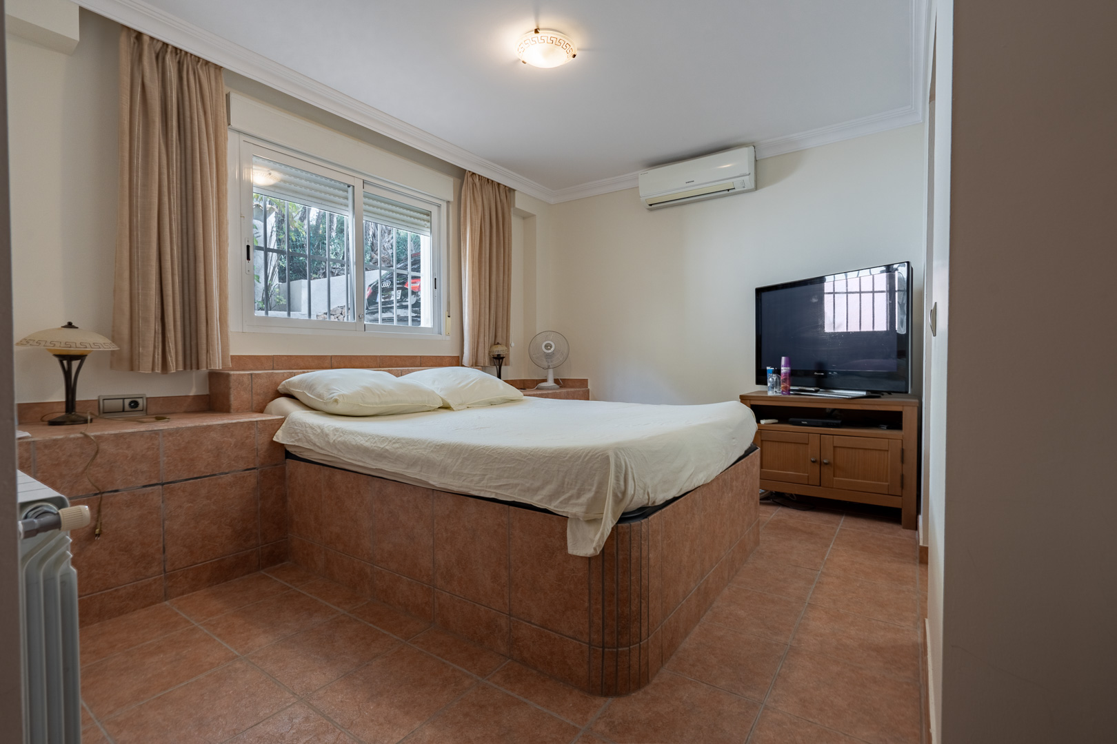 Bedroom featuring a custom tiled bed frame and matching flooring, equipped with split-unit air conditioning, radiator heating, and a television area.