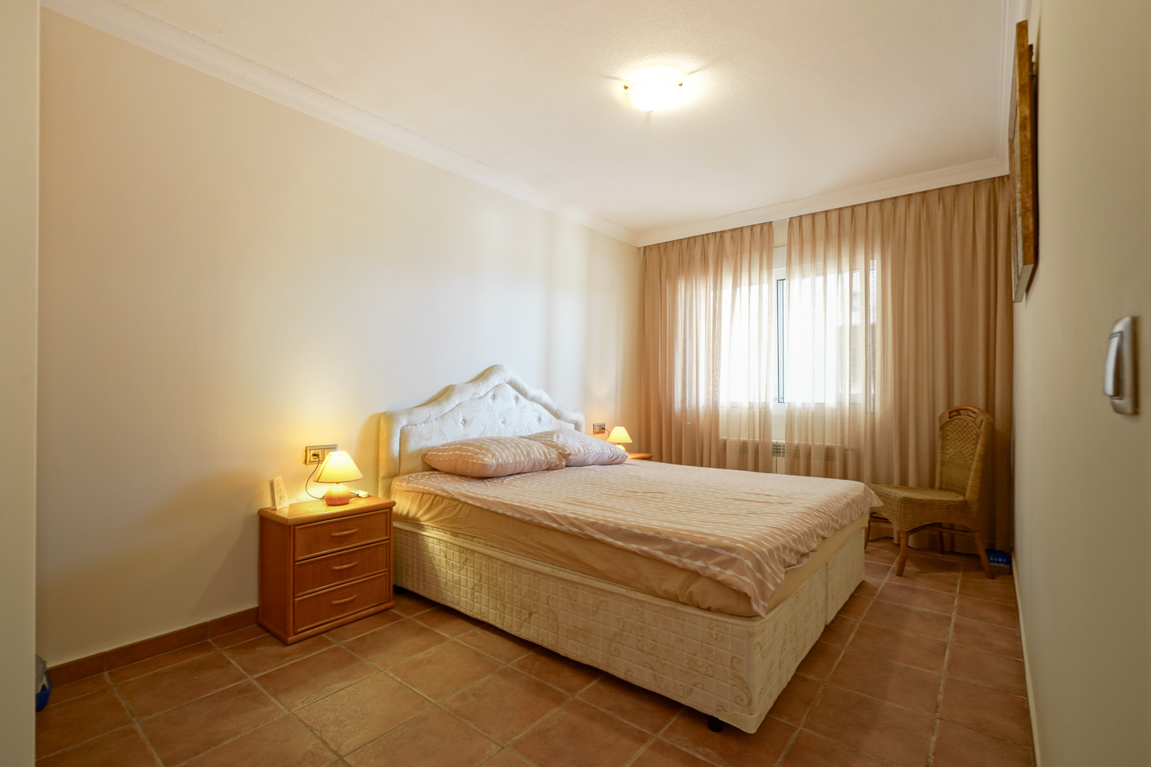 Bedroom featuring terracotta tile flooring, a double bed with a tufted headboard, wooden bedside tables, and a large window providing soft natural light.