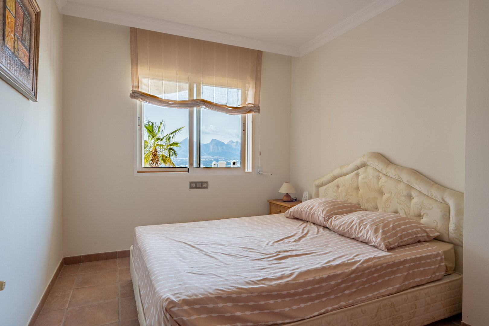 Bedroom featuring terracotta tile flooring and a tufted headboard. The window frames a view of the mountain range and a palm tree in the garden.