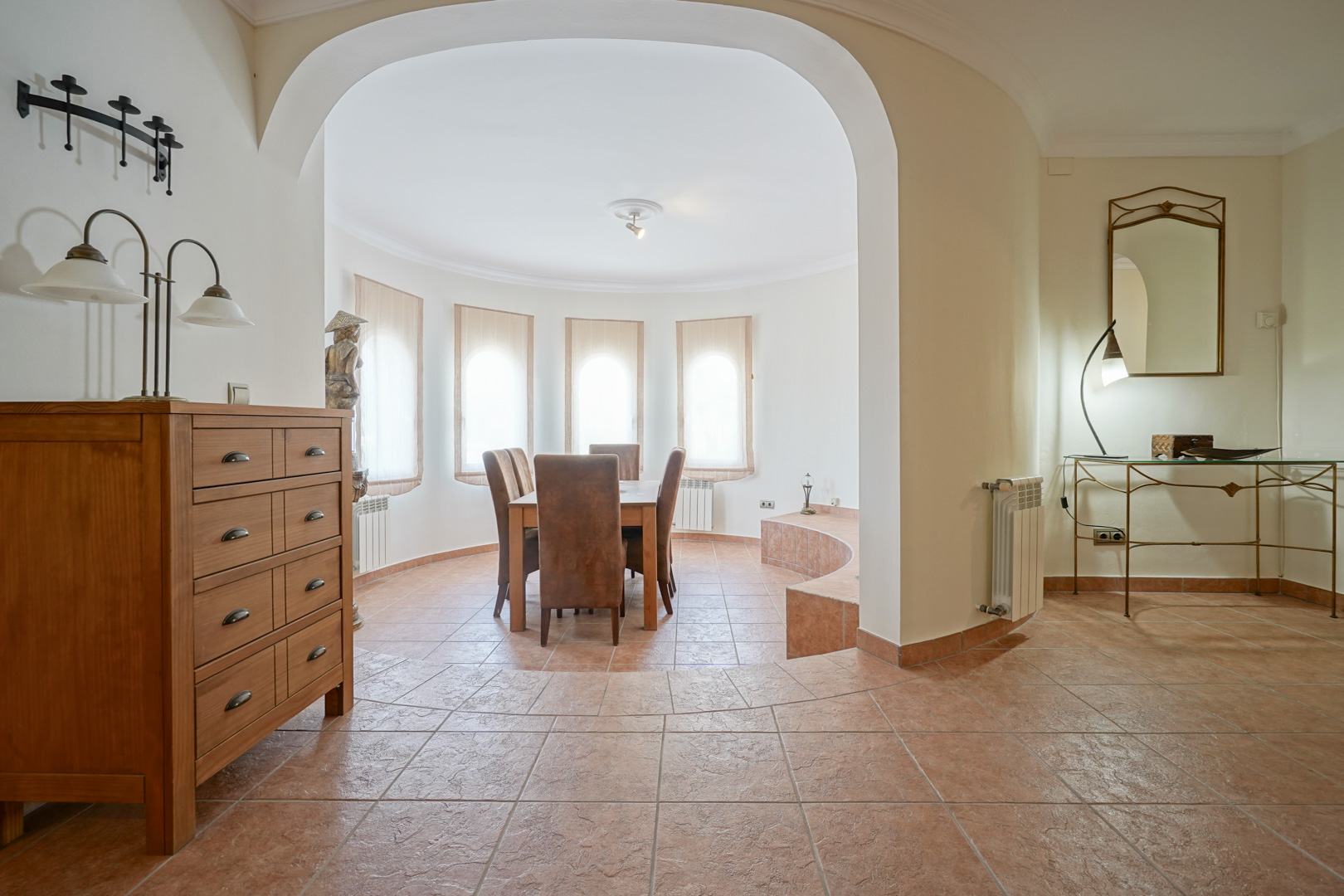 Bright dining area featuring arched windows and terracotta tile flooring. The circular architectural layout includes a wooden dining set and seamless access from the hall.