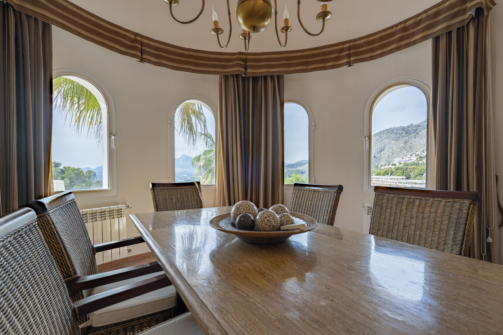 Circular dining area featuring a polished stone table and rattan chairs. Four arched windows provide panoramic views of the Sierra de Bernia mountains and surrounding greenery.
