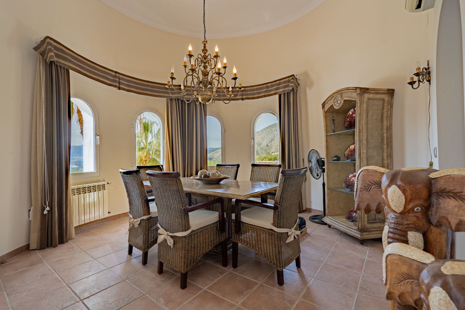 Circular dining area featuring a stone-top table, wicker chairs, and a classic chandelier. Four arched windows provide scenic views of the Sierra de Bernia mountains.
