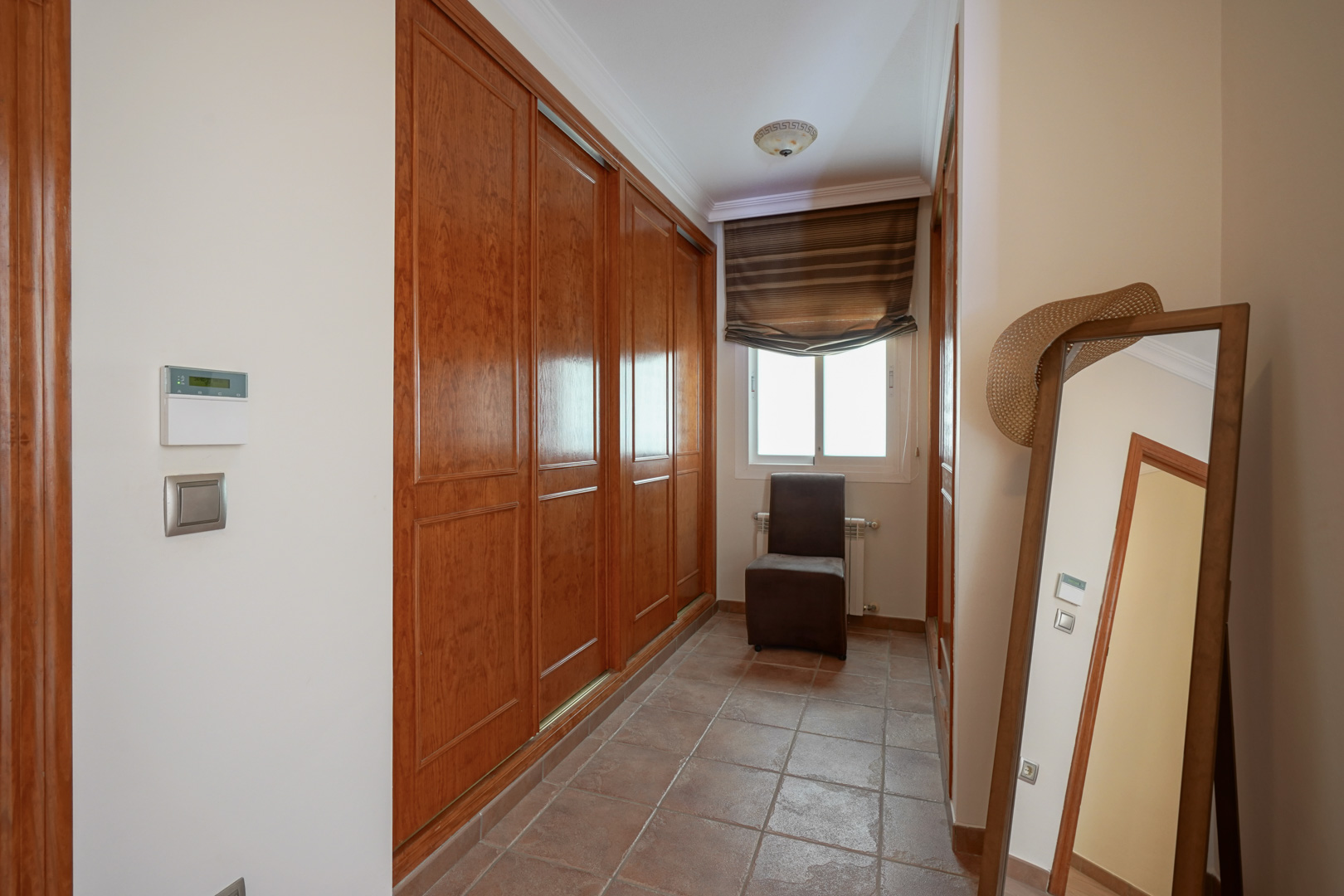 Dressing area featuring large built-in wooden wardrobes, terracotta tile flooring, and a window for natural light. Equipped with a full-length mirror and a side chair.