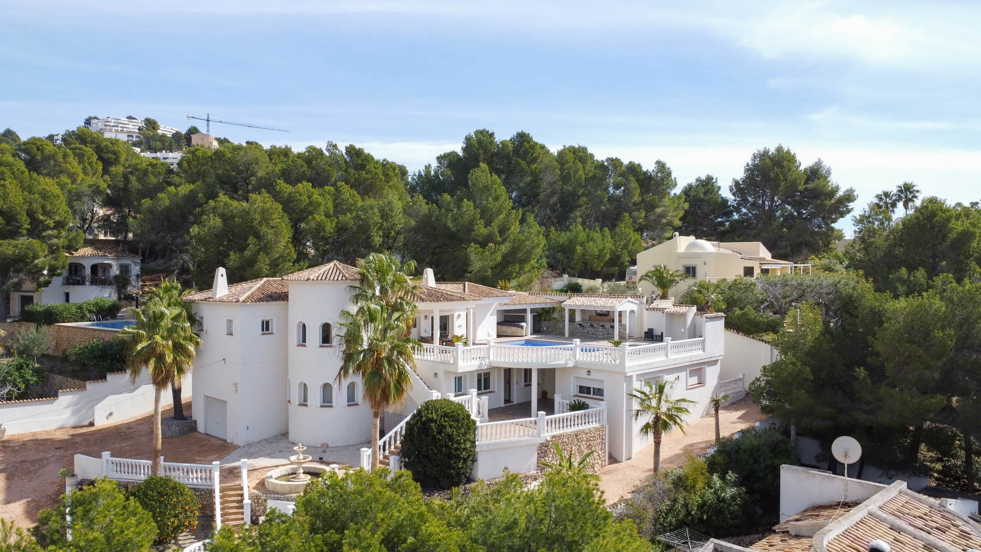 Exterior view of the Mediterranean villa featuring a white facade, circular tower, and multi-level terraces with balustrades, set against a lush pine forest.
