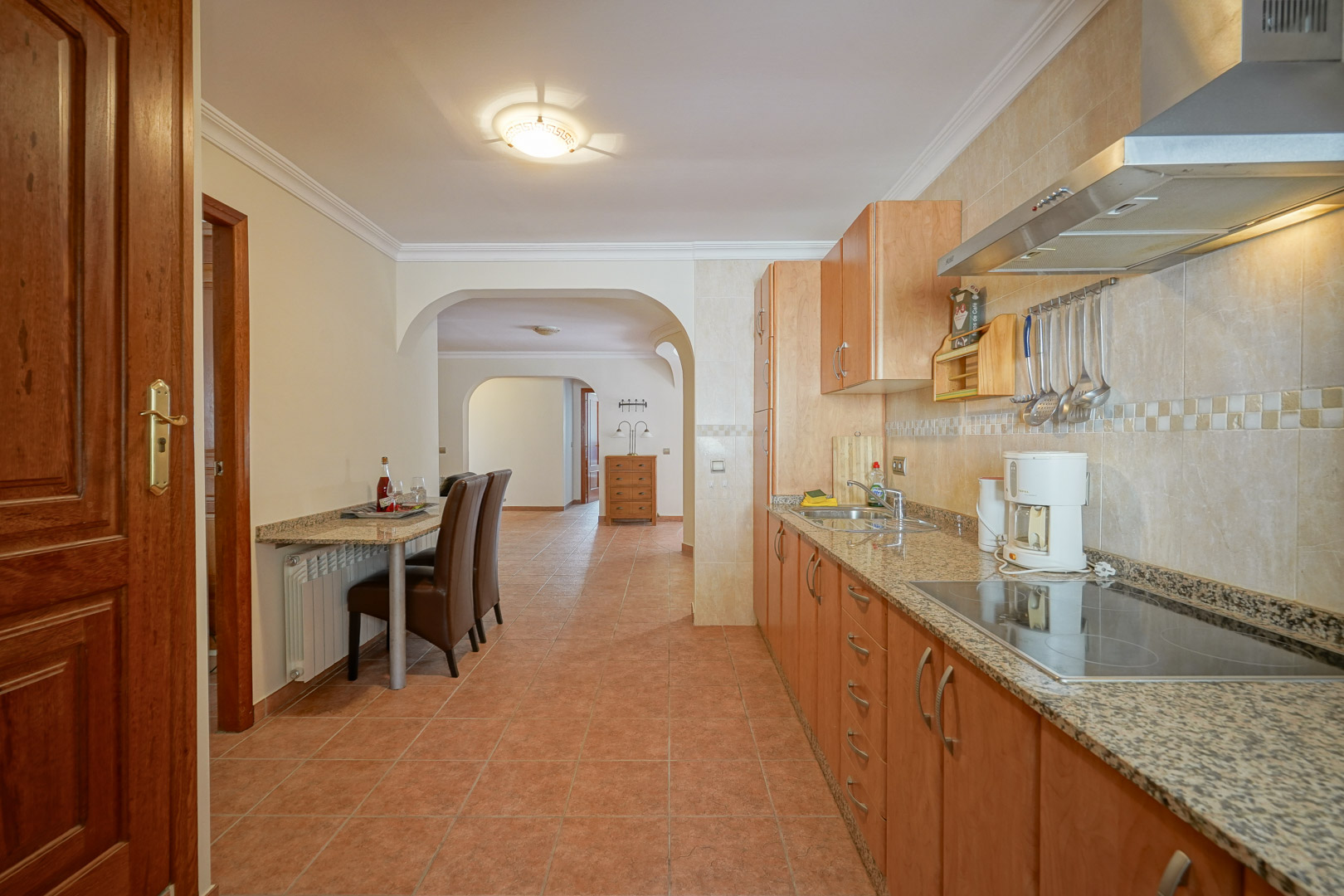 Functional kitchen with wood cabinetry, granite countertops, and a ceramic hob. It features an integrated breakfast bar and tiled flooring, connecting to the living area via elegant arches.