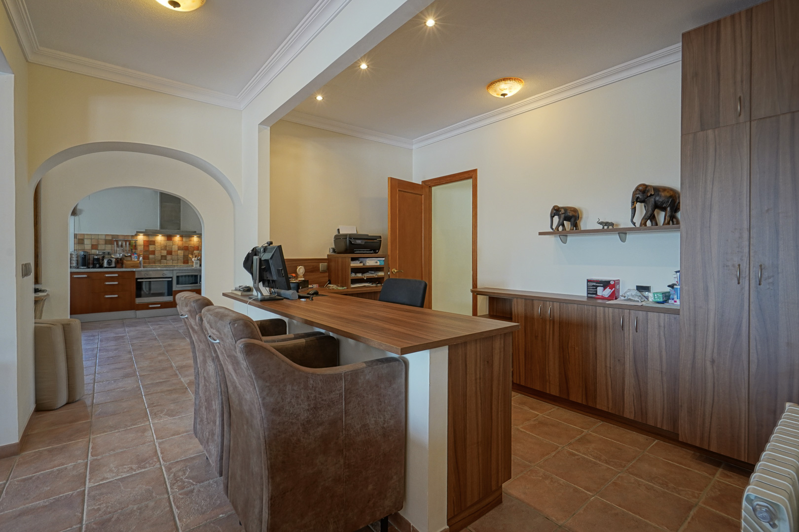 Home office area featuring custom wood cabinetry, terracotta tile flooring, and arched entryways leading to the kitchen.