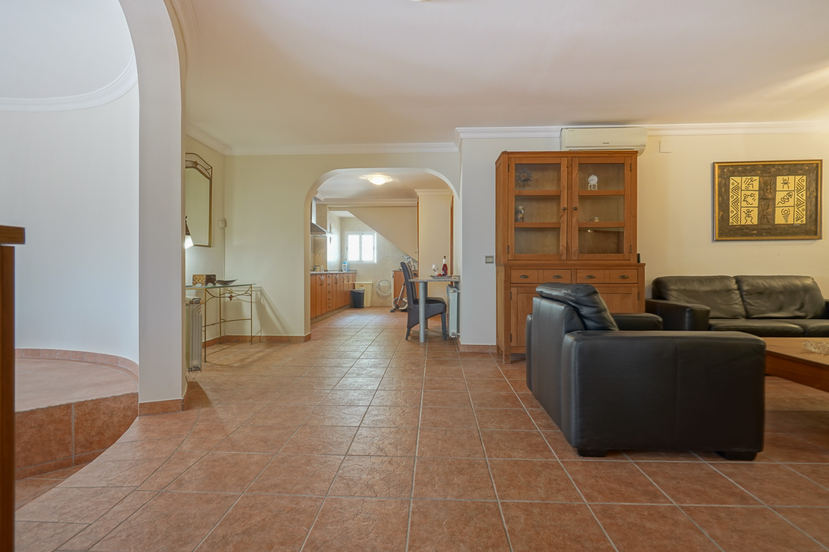Living area featuring black leather seating and terracotta-style tiling, with an arched opening leading to a wooden-cabinet kitchen and dining nook.