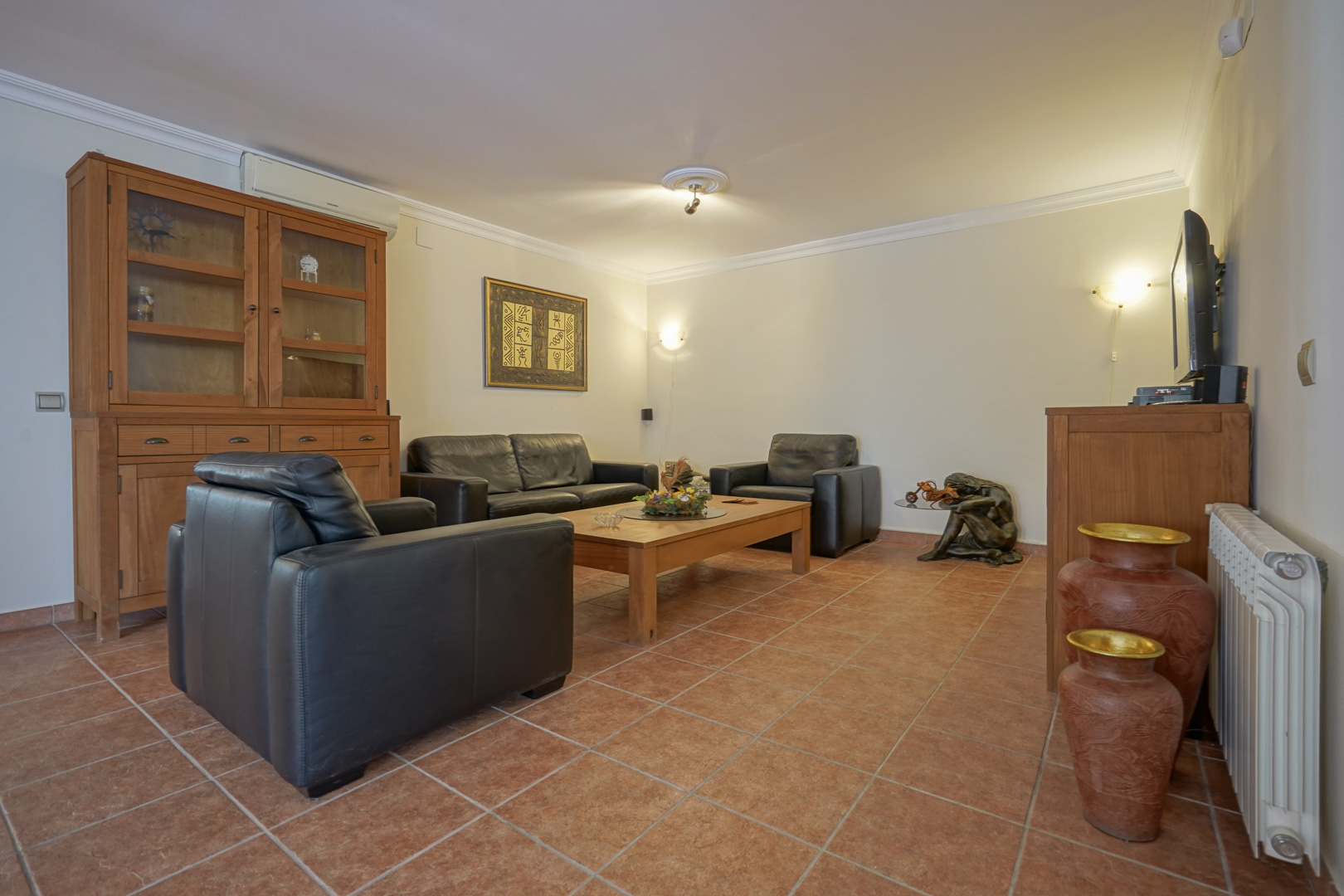 Living area featuring black leather seating, solid wood furniture, and terracotta tile flooring. Equipped with split air conditioning and central heating radiators.
