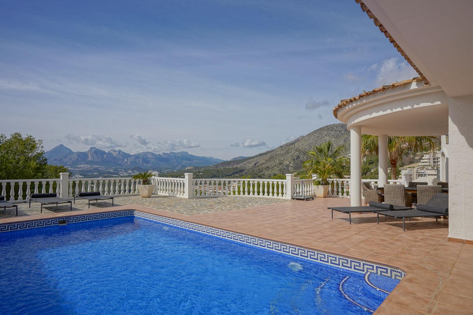 Main terrace featuring a blue tiled pool with a Greek key border, sun loungers, and a covered porch. It offers panoramic views of the Puig Campana and Sierra Aitana.