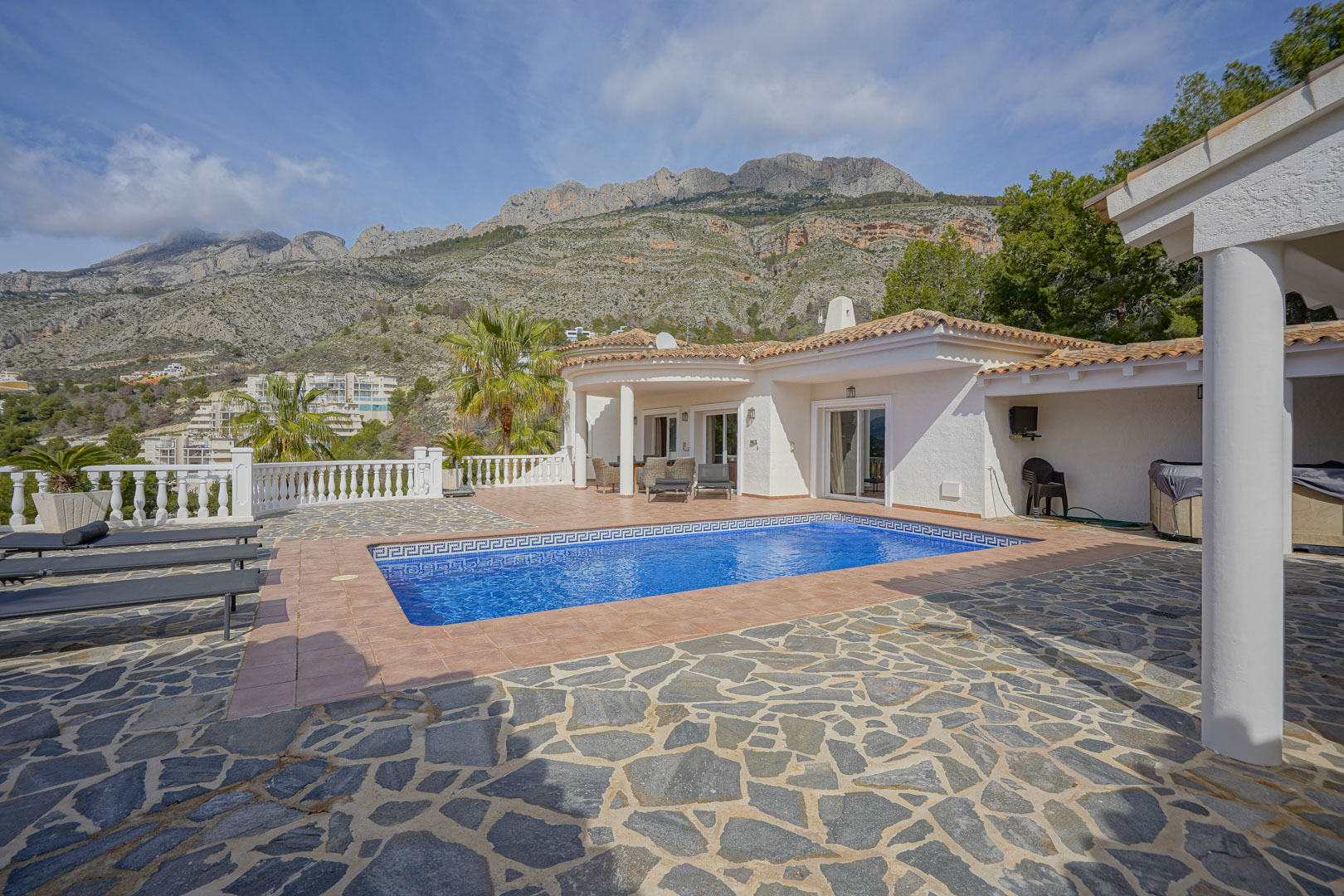 Main terrace featuring a rectangular pool with a mosaic border and views of the Sierra de Bernia. The area includes natural stone paving, terracotta tiles, and a white balustrade.