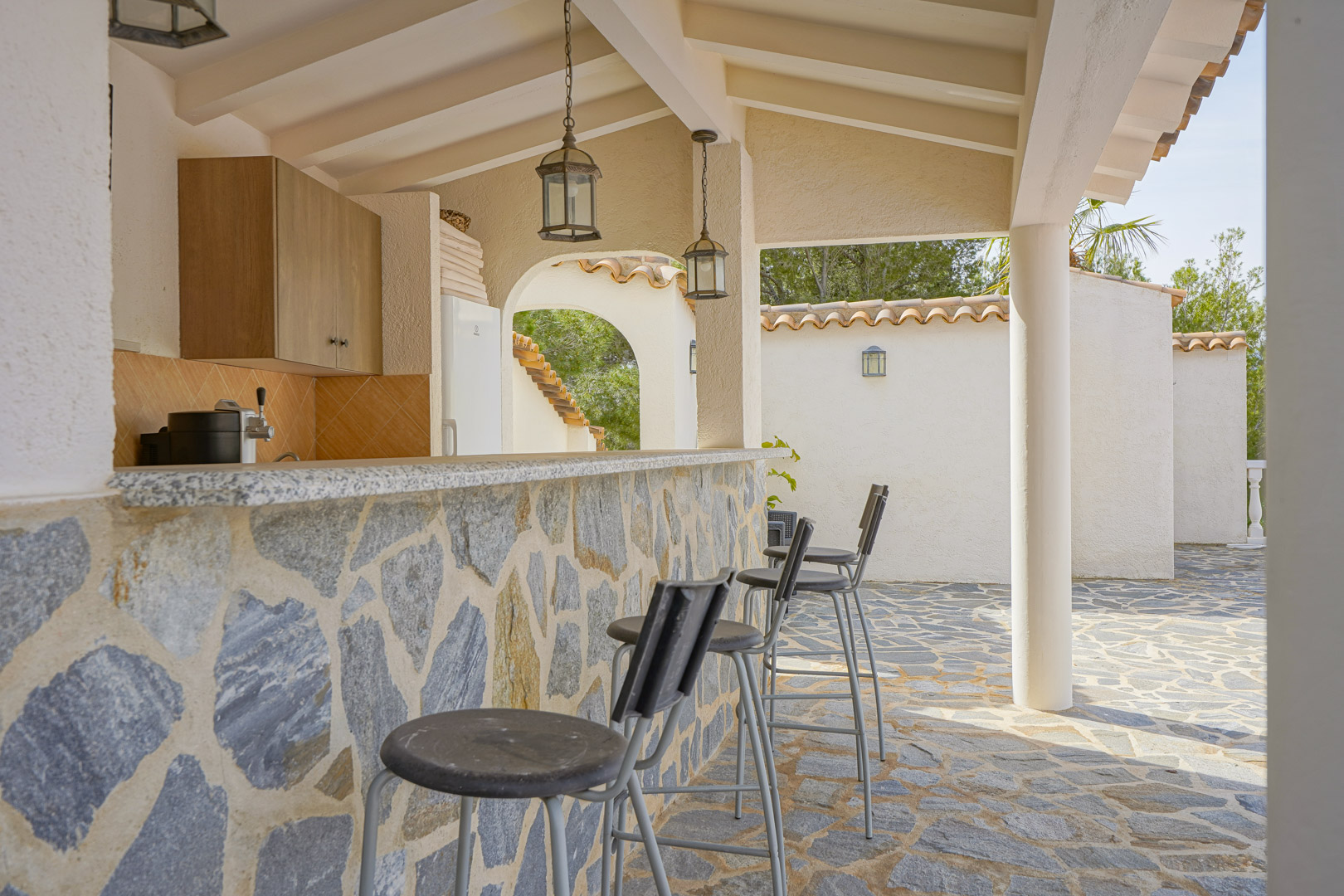 Outdoor kitchen and bar featuring natural stone cladding, granite countertops, and high stools under a beamed porch ceiling with rustic pendant lanterns.