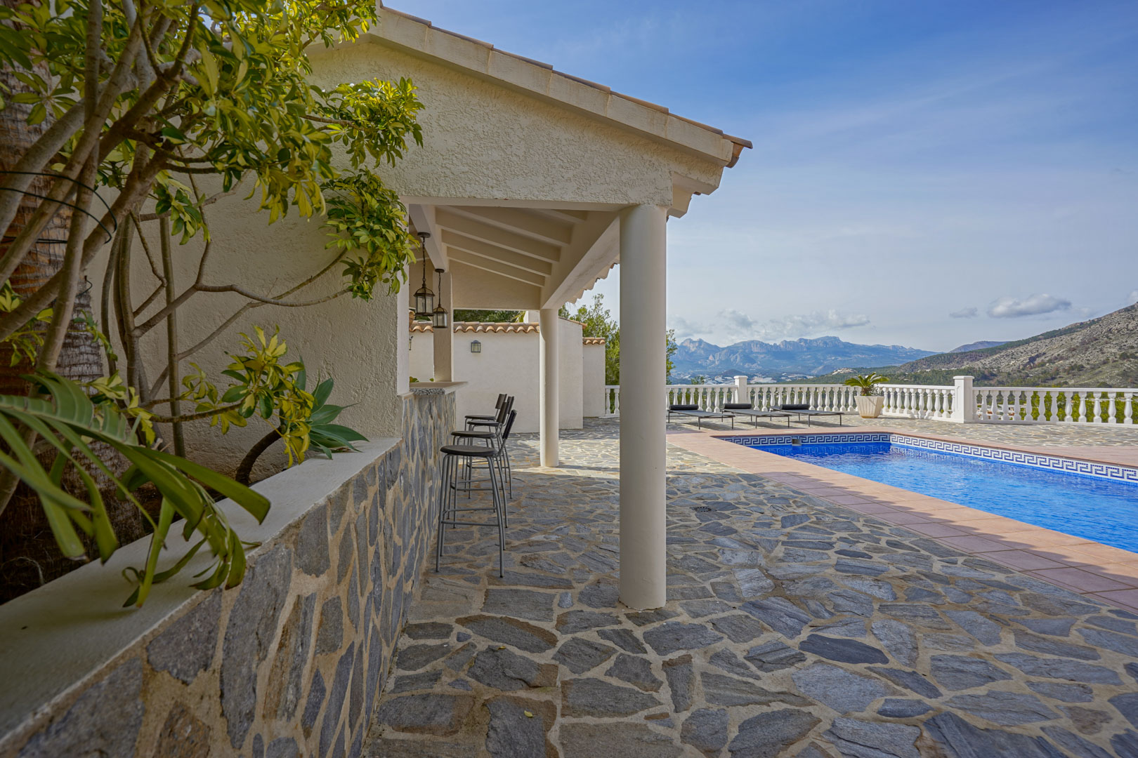 Outdoor terrace featuring natural stone flooring, a covered summer kitchen with bar seating, and panoramic mountain views beside the private pool.