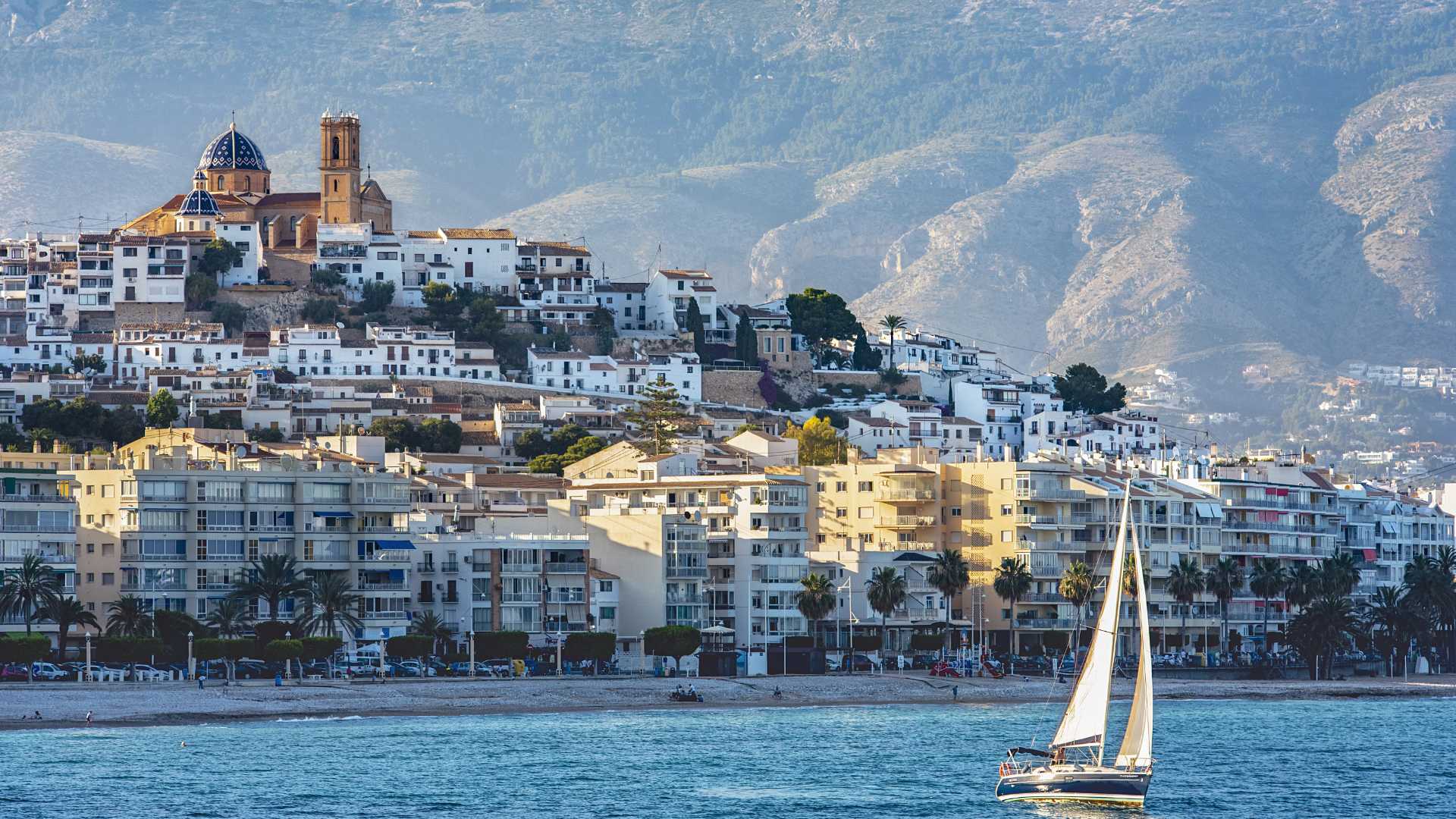 Panoramic view of Altea's old town featuring the iconic blue-domed church, set against the Sierra de Bernia mountains with a sailboat on the Mediterranean.