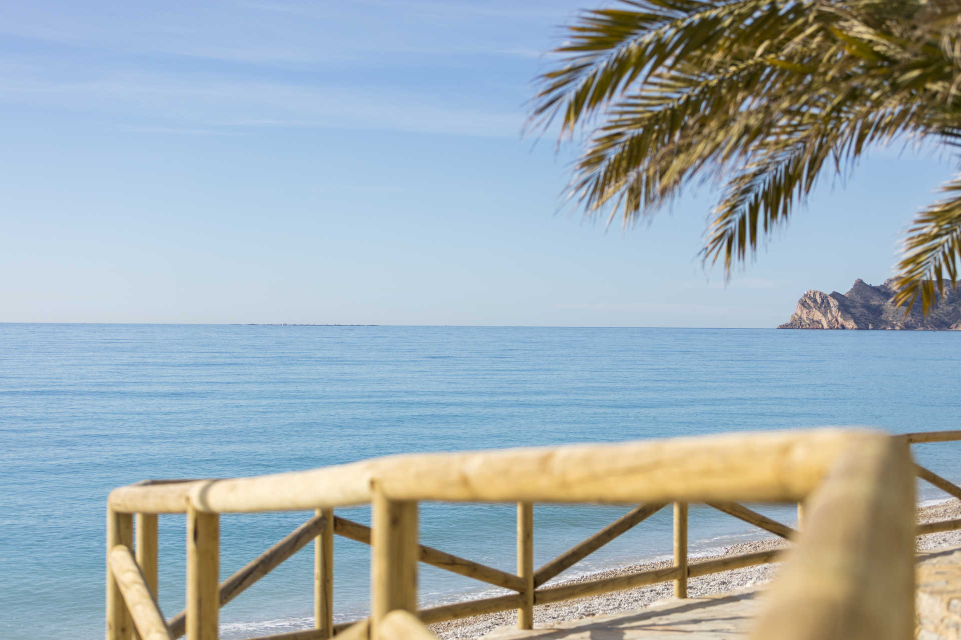 Panoramic view of the Mediterranean Sea from Altea's coastline, featuring wooden railings, a pebble beach, and distant rocky cliffs under a clear blue sky.