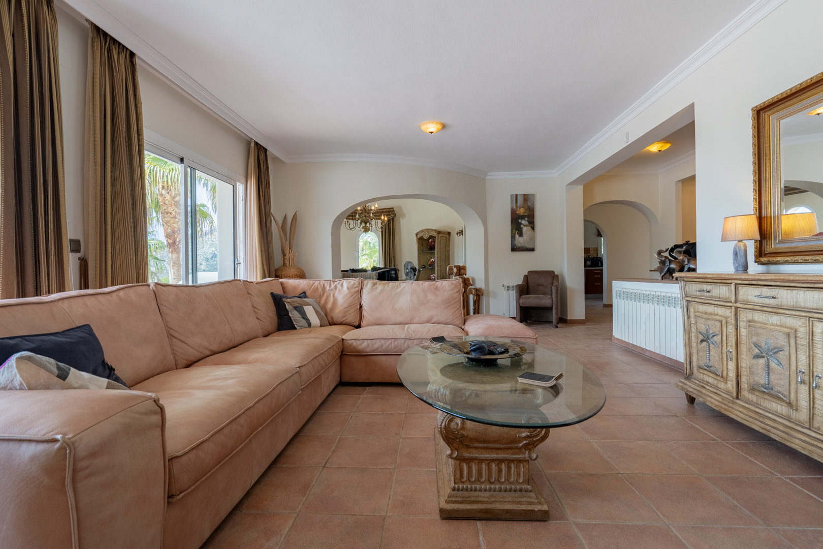Spacious living area with terracotta tile flooring, a large L-shaped leather sofa, and arched entryways. A glass-topped coffee table sits near a carved wooden sideboard.